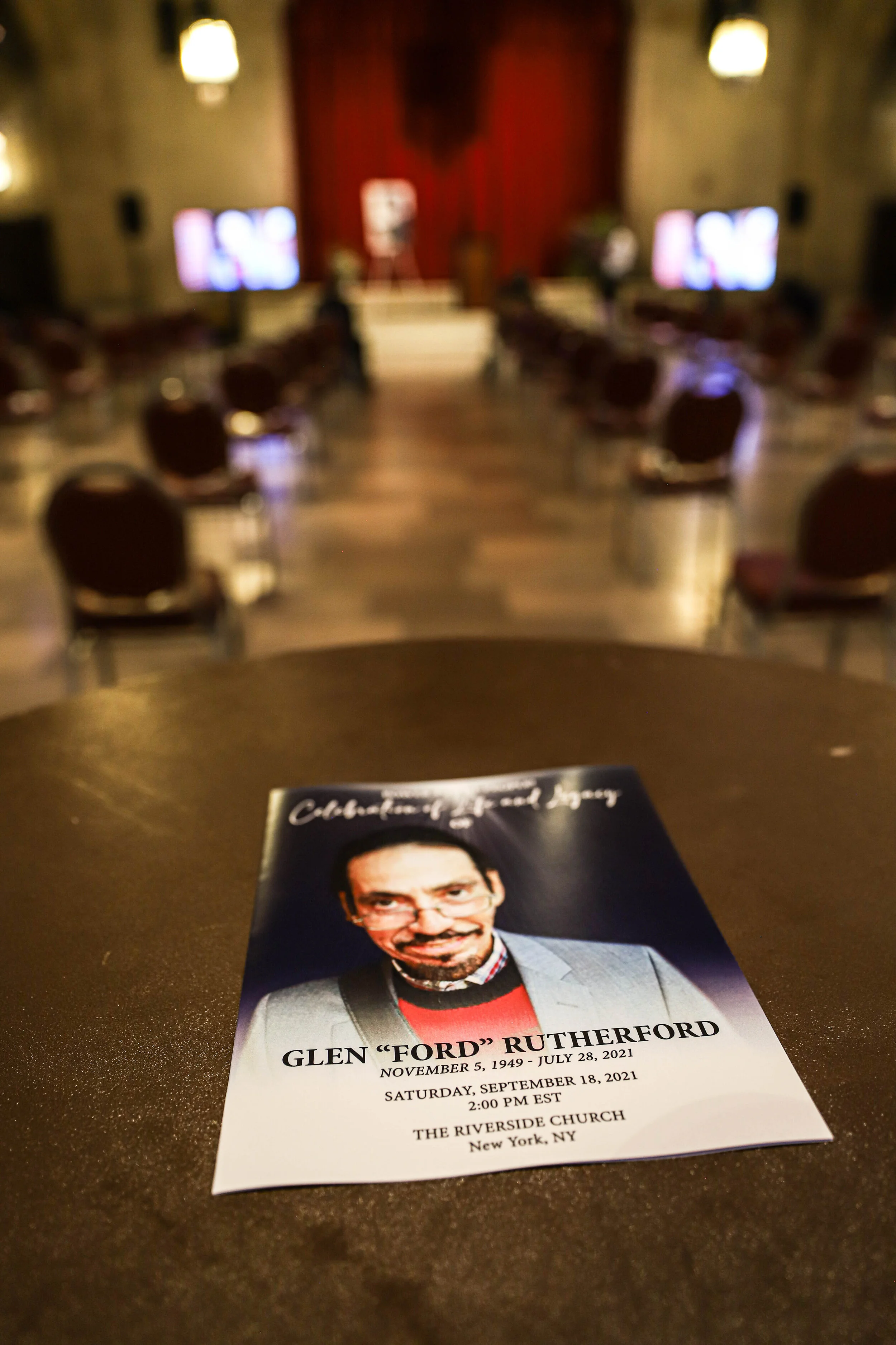 A leaflet on a dark table with a photograph of Glenn "Ford" Rutherford and details of a memorial service. In the background, a blurred view of an indoor space with chairs and television screens.