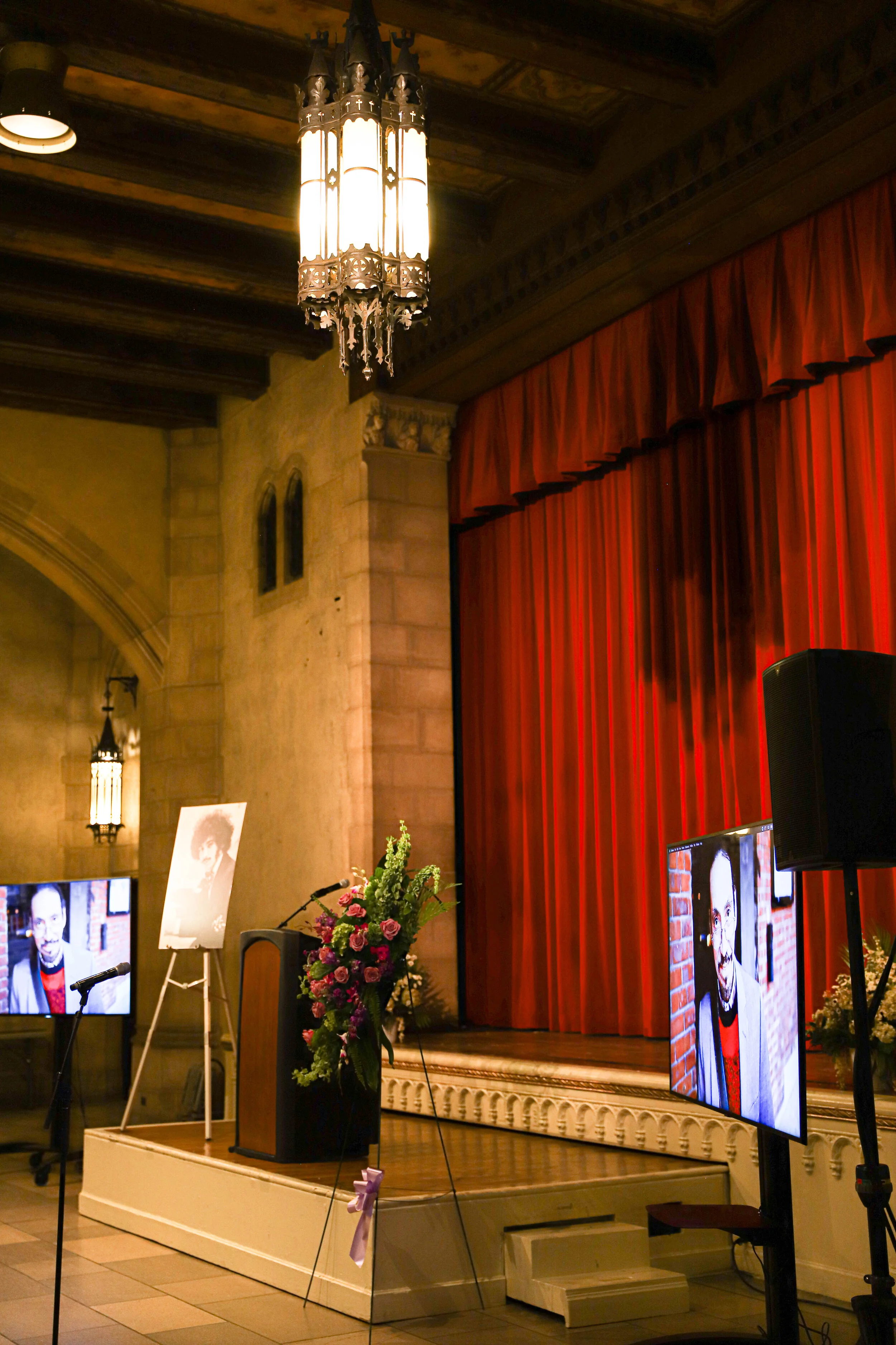 An indoor stage with red curtains, a floral arrangement at a podium, and multiple screens displaying images of a person. The room has stone walls, wooden beams on the ceiling, chandeliers, and decorative lighting.