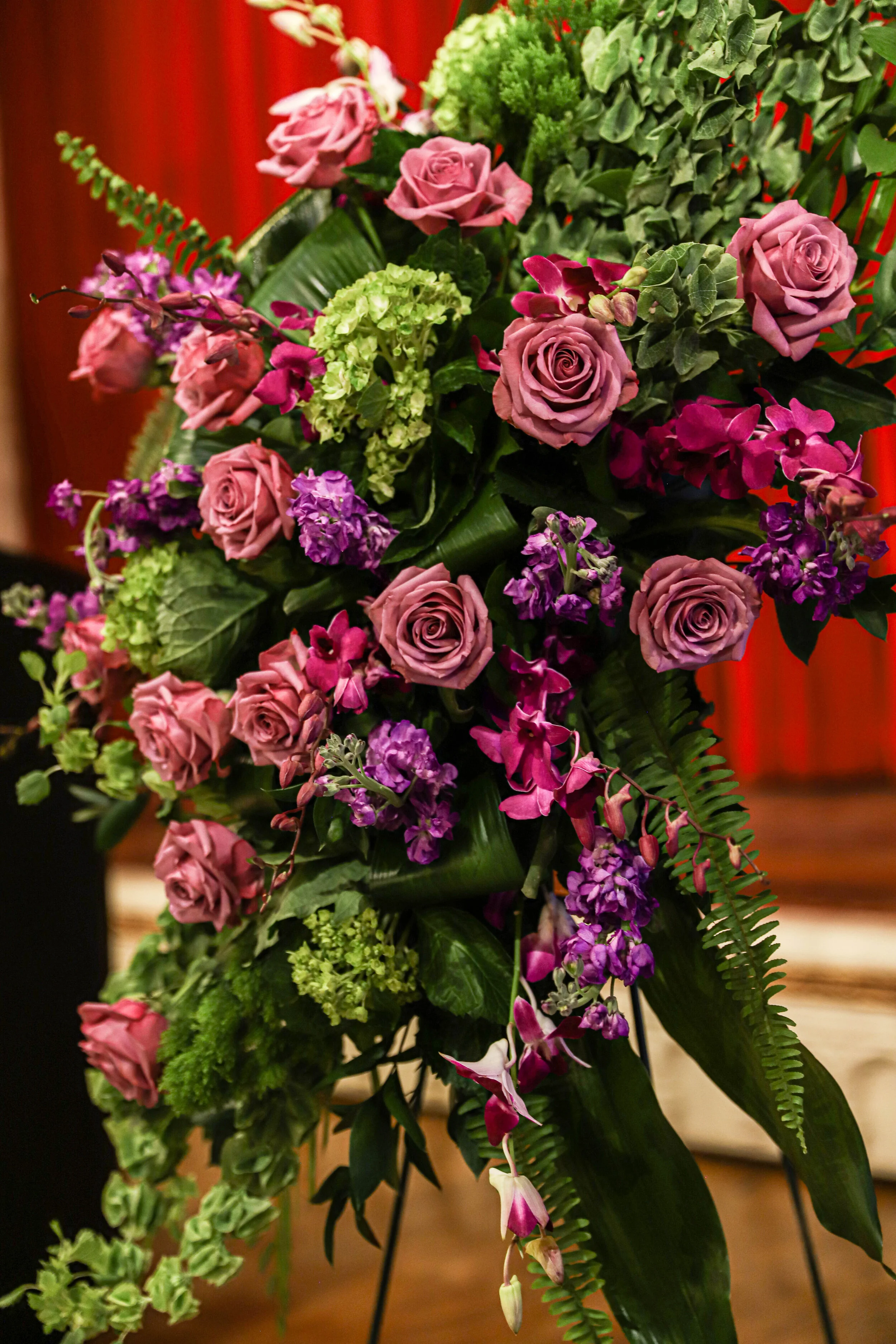 A lush arrangement of pink roses, purple orchids, green hydrangeas, and various green foliage, including ferns, arranged in a bouquet and set against a warm wooden background.