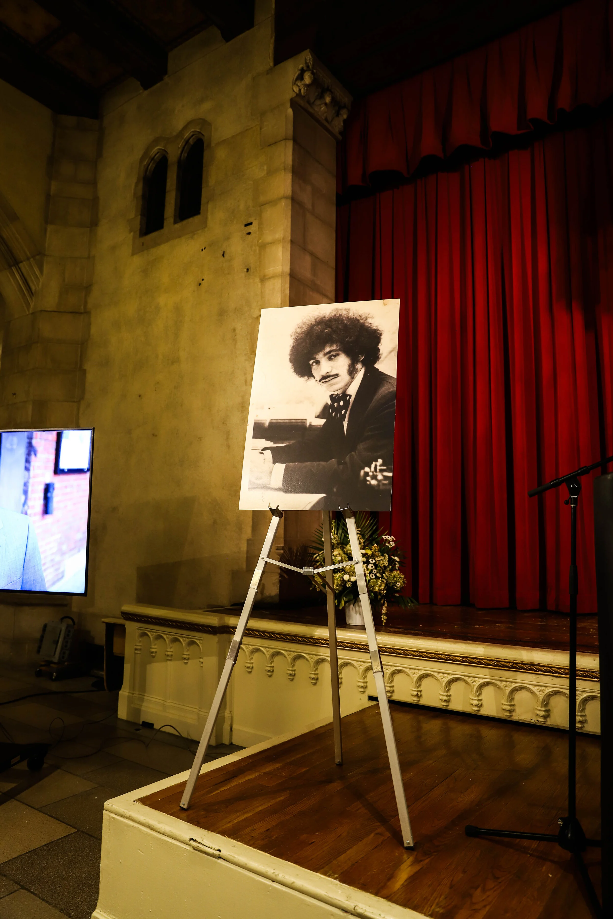 A stage with red curtains, a large black-and-white poster of a man with curly hair playing the piano, and a TV screen displaying a blurred image of a person in the background. The stage has a wooden floor, and there are flowers in front of the poster