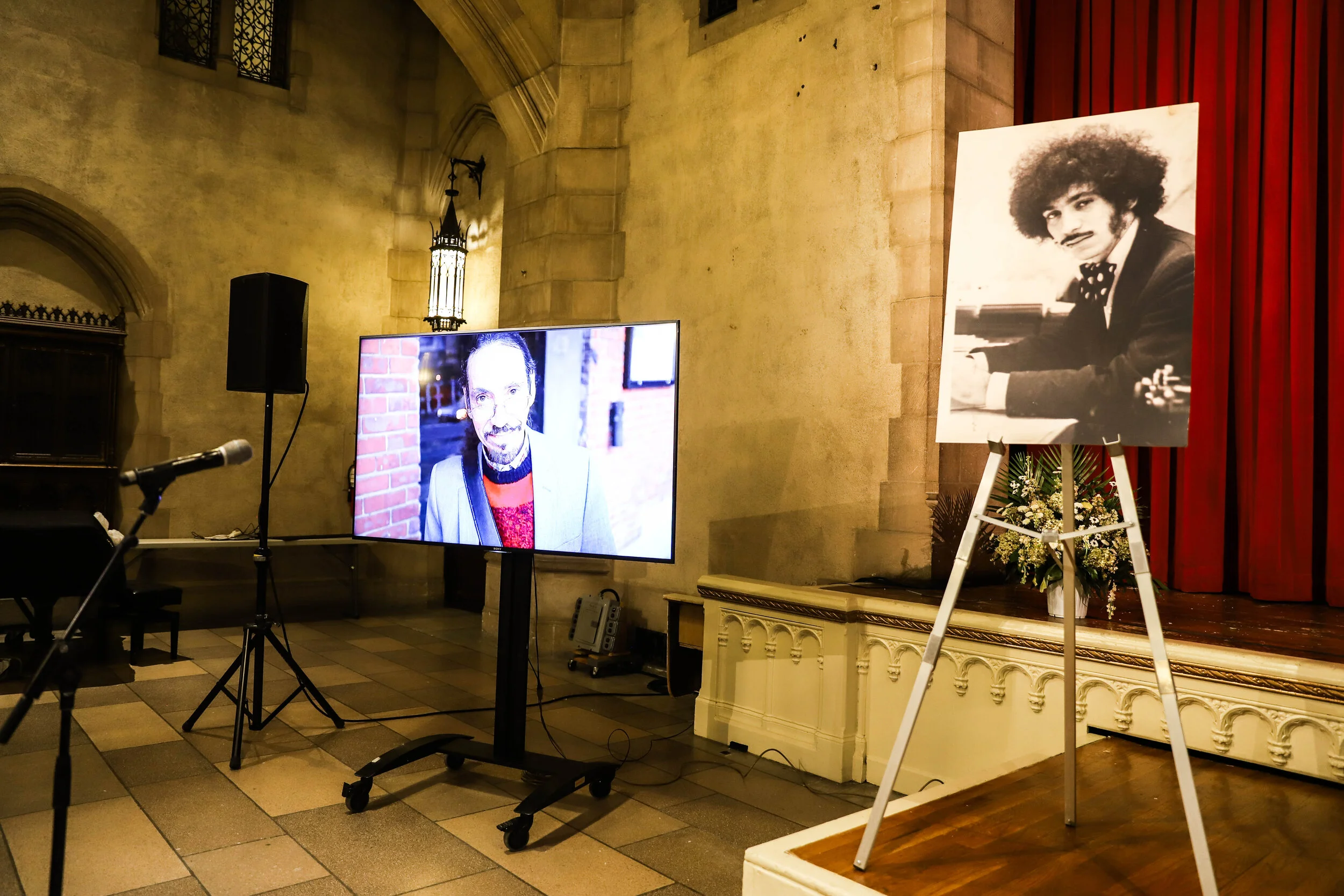A memorial setup with a large black-and-white portrait on an easel of a man with curly hair and facial hair, wearing a suit and bowtie, and a floral arrangement at the base. There are monitors, a microphone, and speakers in a room with stone walls an