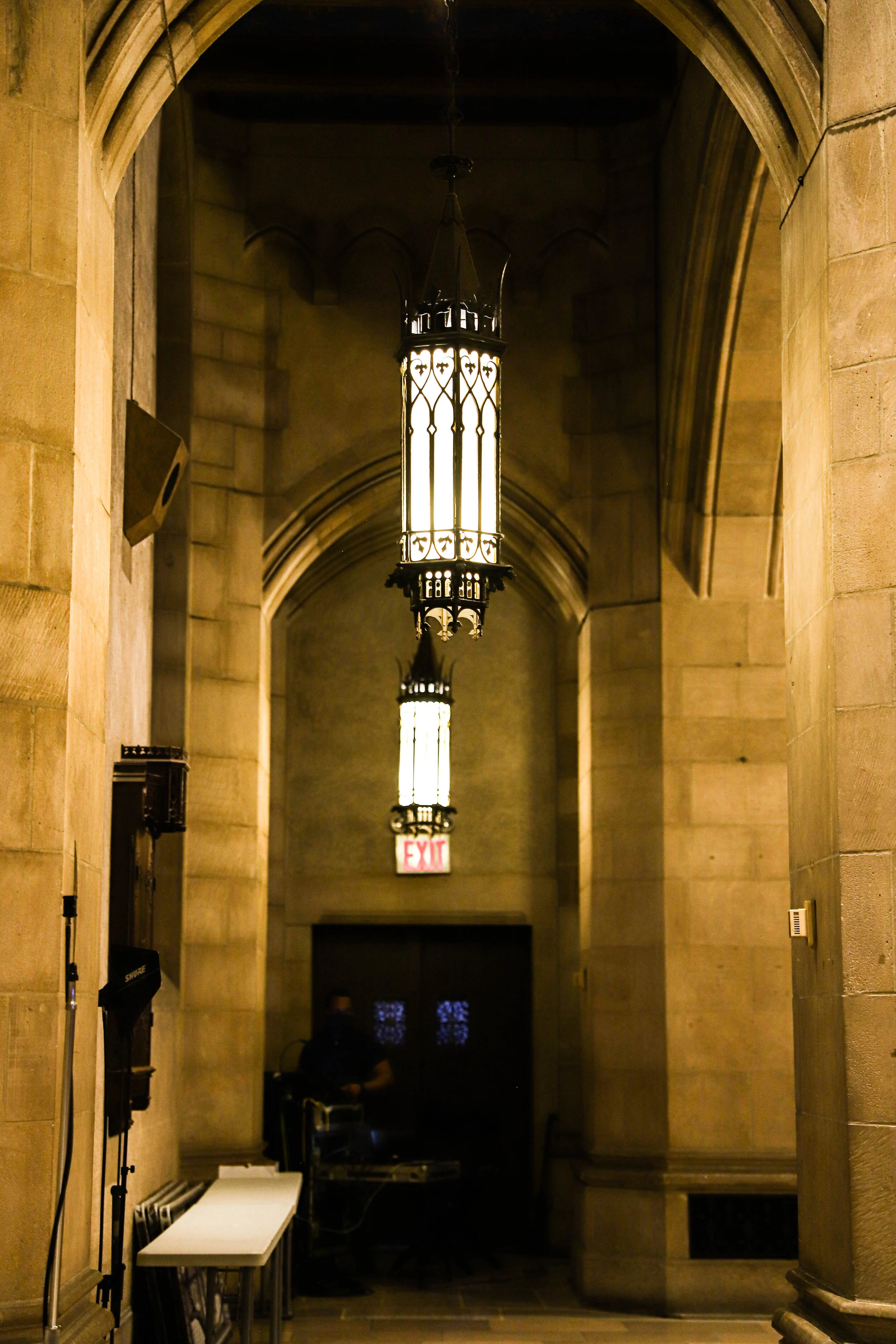 A dimly lit hallway with arched ceilings and hanging ornate lantern-style lights. There is a white table with chairs on the left side and an exit sign illuminated in red at the end of the hallway.