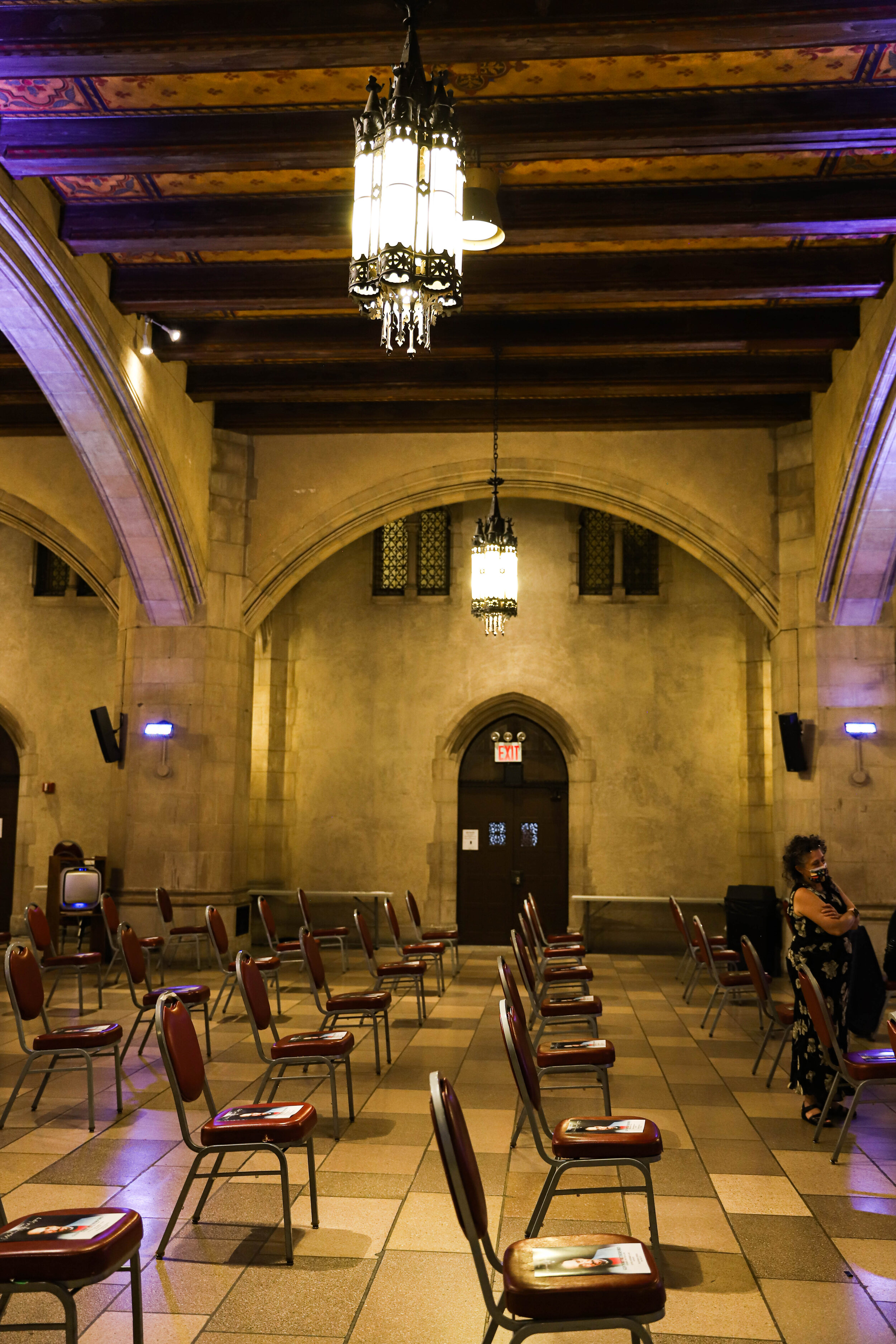 Interior of a large hall with chairs arranged in rows, lit chandeliers hanging from a decorated wooden ceiling, arched windows, and a woman wearing a black and white floral dress standing on the right side.