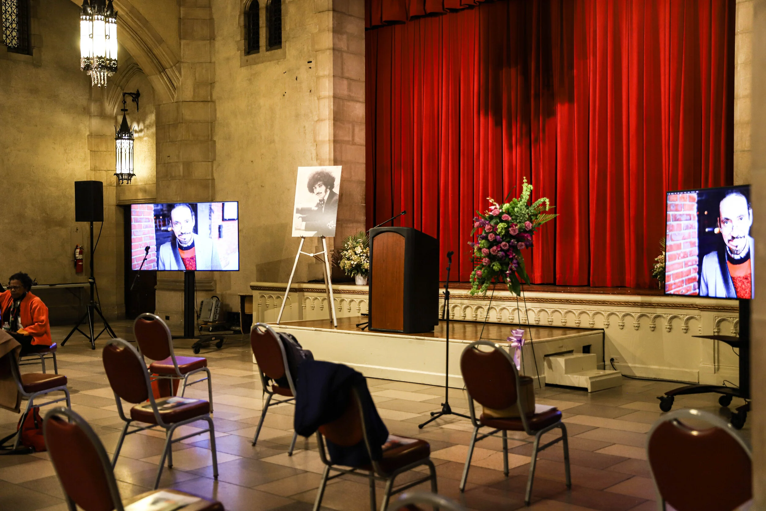 An indoor event space with a stage, red velvet curtains, and a large floral arrangement on the right side of the stage. There are two television screens displaying a woman with dark hair and a black blazer, against a brick wall. The room has stone wa