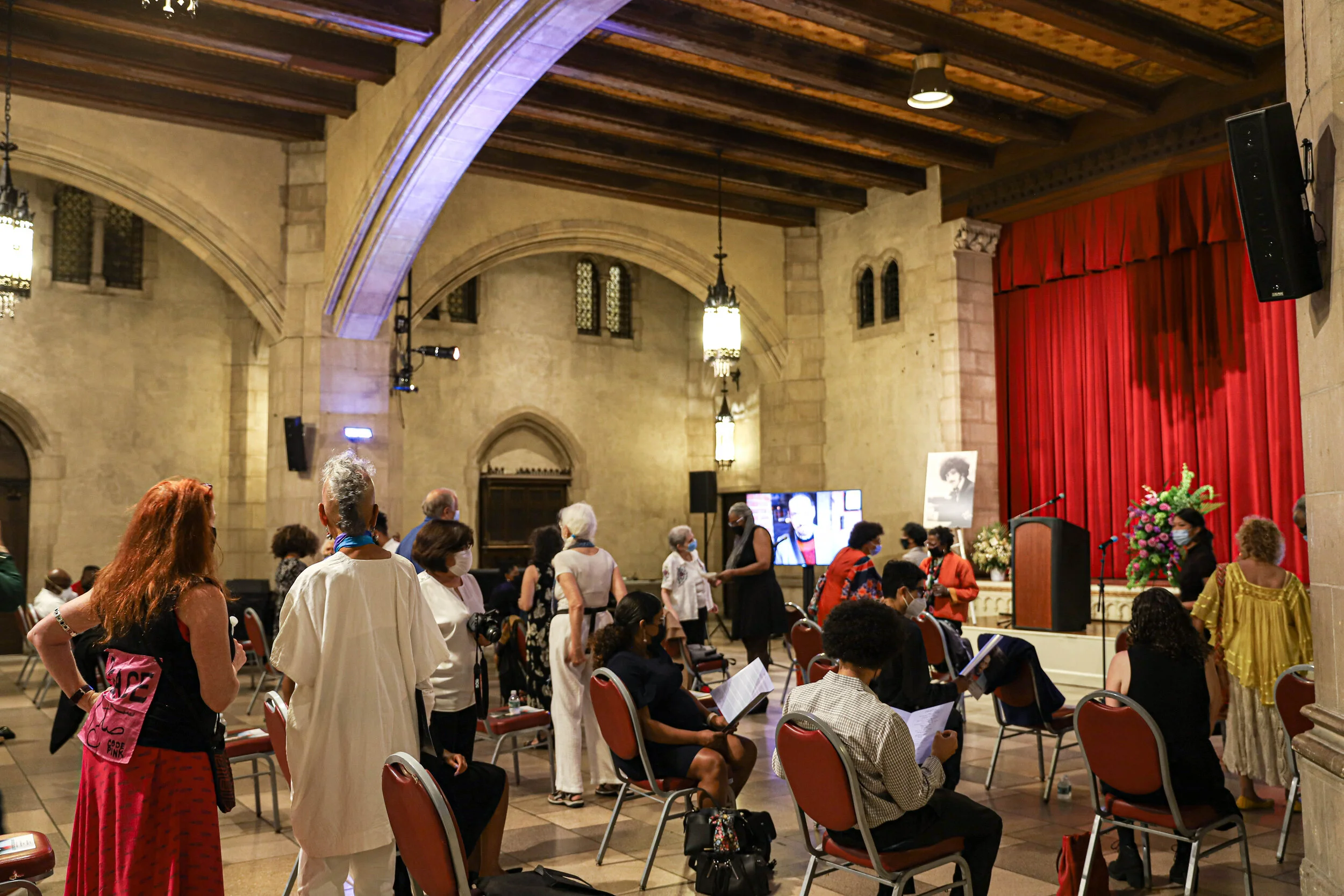 People gathered inside a large hall with stone walls, wooden ceiling beams, and stained glass windows. Some individuals are standing, sitting, and wearing masks. A stage with a red curtain and floral arrangements is visible, along with a television s