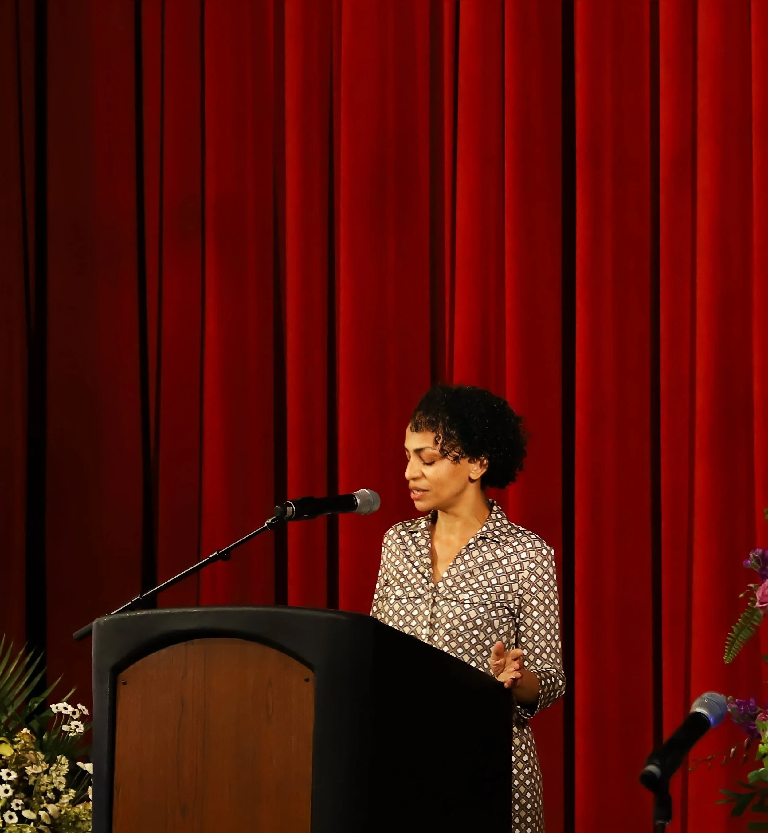 A woman with short curly hair, wearing a patterned dress, speaking at a podium with a microphone in front of a red curtain backdrop. There are flowers and plants visible on the side.