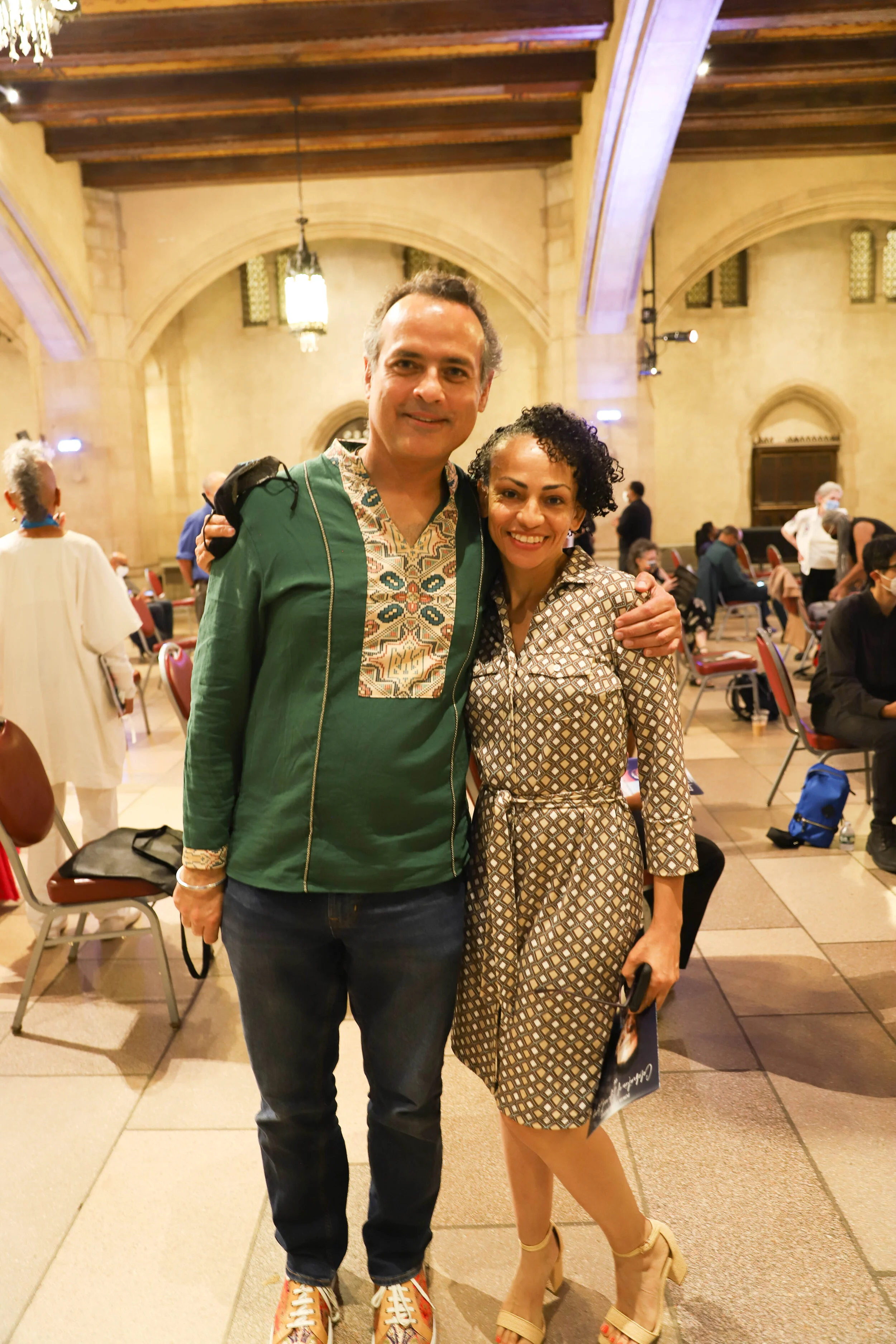 A man and woman standing close together inside a large, ornate building, smiling at the camera. The man is wearing a green shirt with colorful embroidery and jeans, while the woman is wearing a patterned dress and high heels. There are other people s
