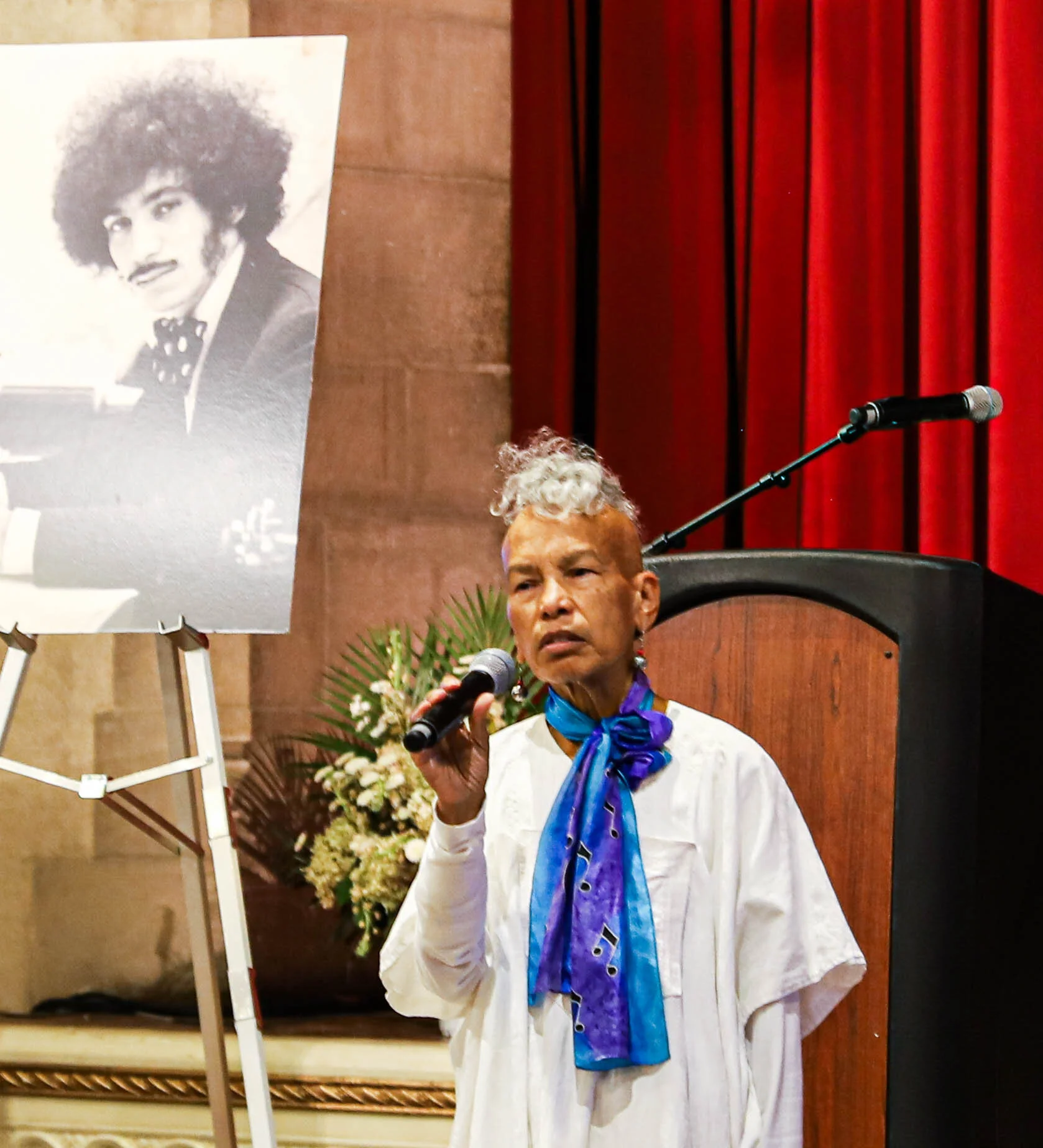 A woman with gray hair, wearing a white outfit and blue scarf, speaking into a microphone at an event. Behind her is a large portrait of a person with curly hair, and there are red curtains and floral arrangements in the background.