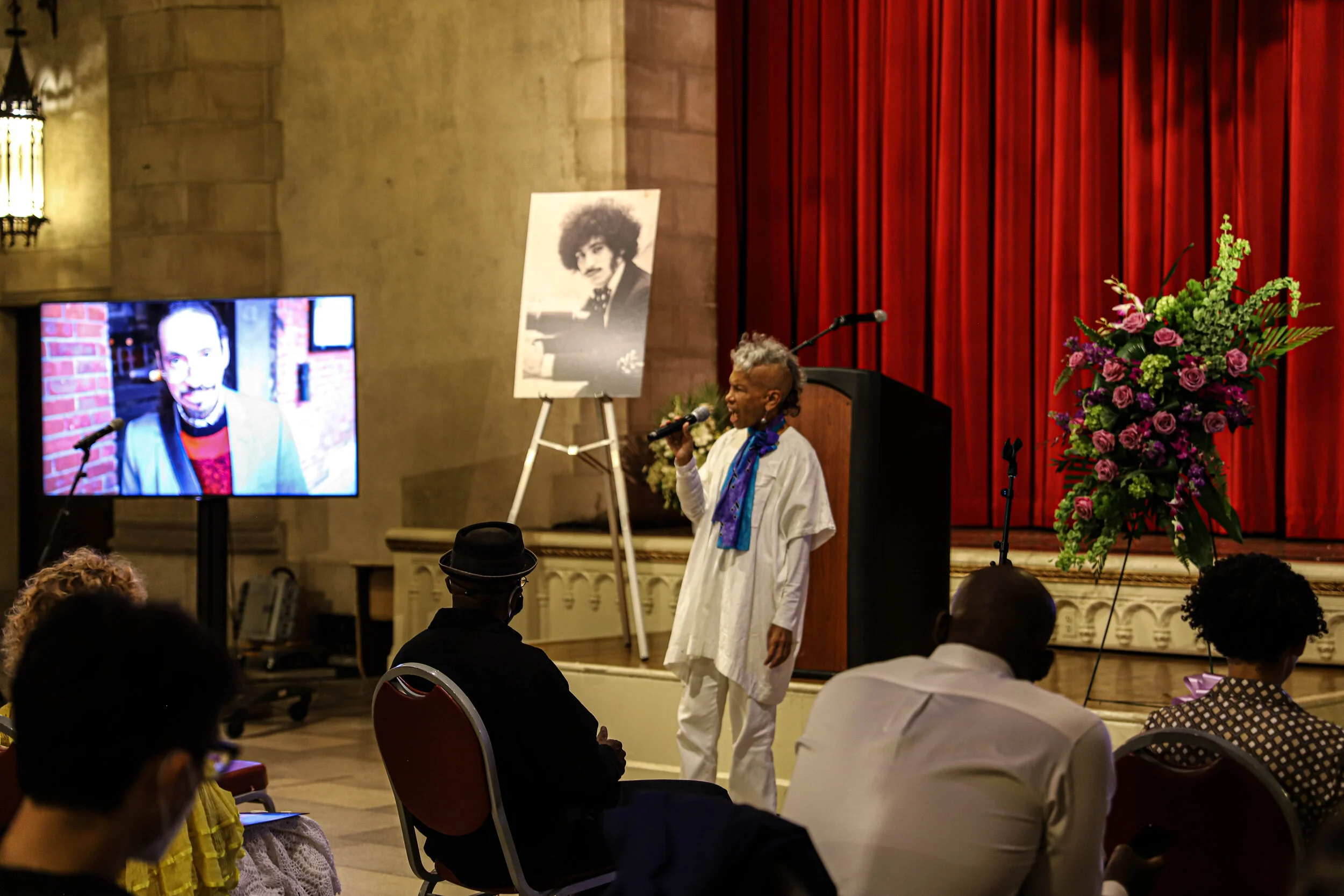 An indoor event on a stage with red curtains, a woman speaking into a microphone, a large floral arrangement on the right, a portrait photograph on a stand, a video screen displaying a man speaking, and audience members seated.