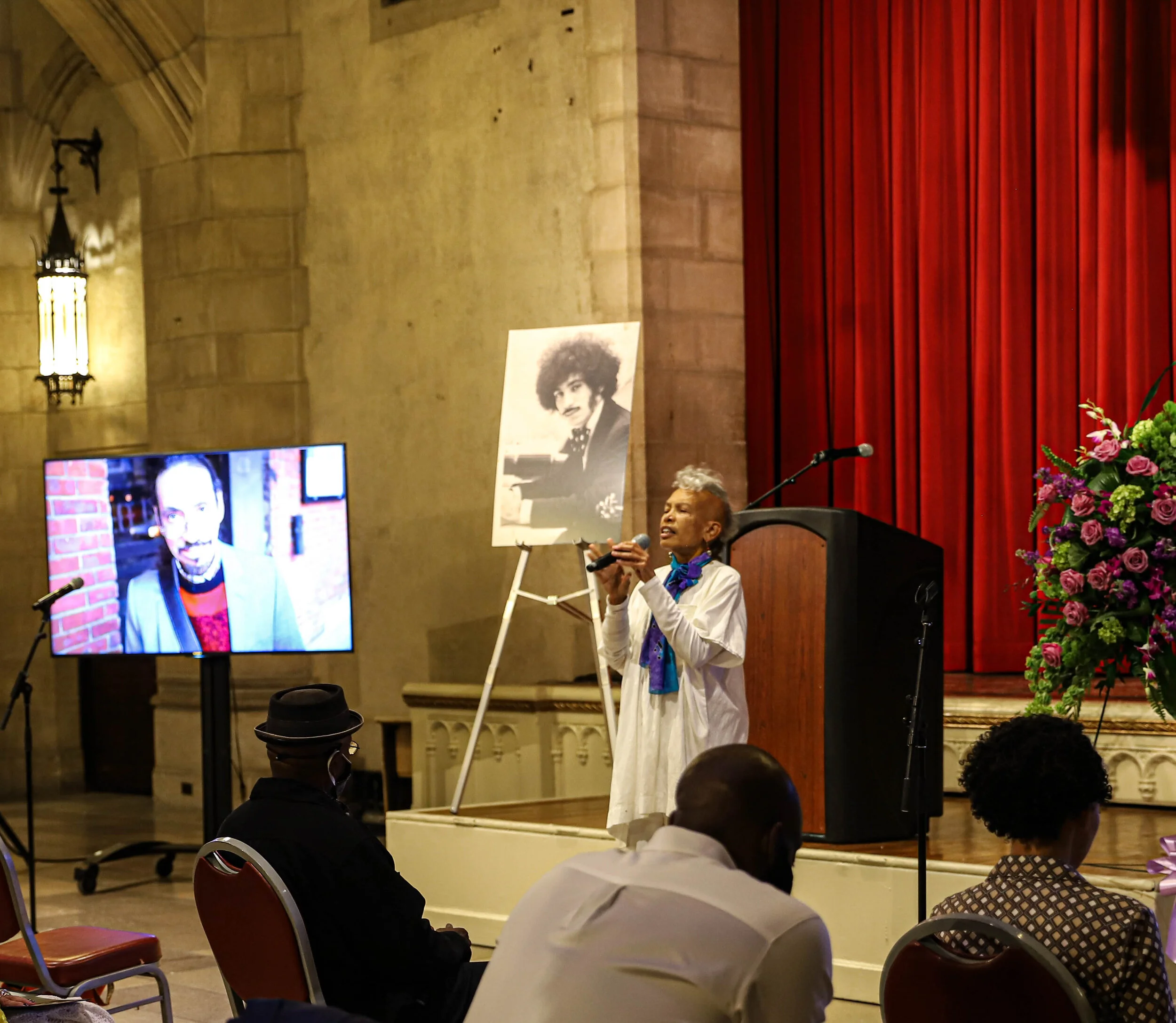 A woman in a white blazer and colorful scarf presenting at a stage with a microphone, holding a large black and white portrait of a woman with curly hair. Audience members are seated facing her in a historic hall with stone walls, red curtains, and f