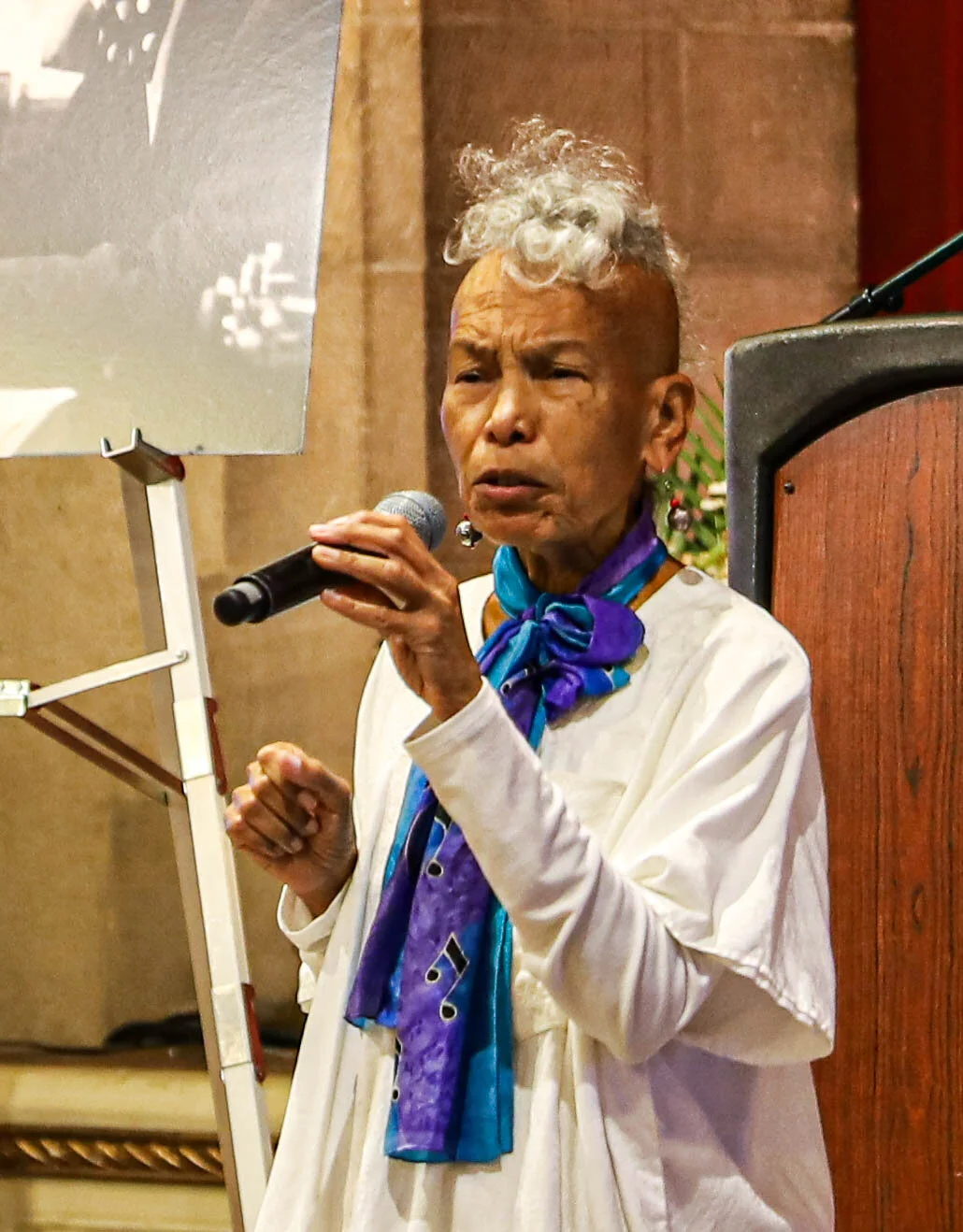 An elderly person with a unique hairstyle holds a microphone and speaks at an event, wearing a white outfit and a colorful scarf.