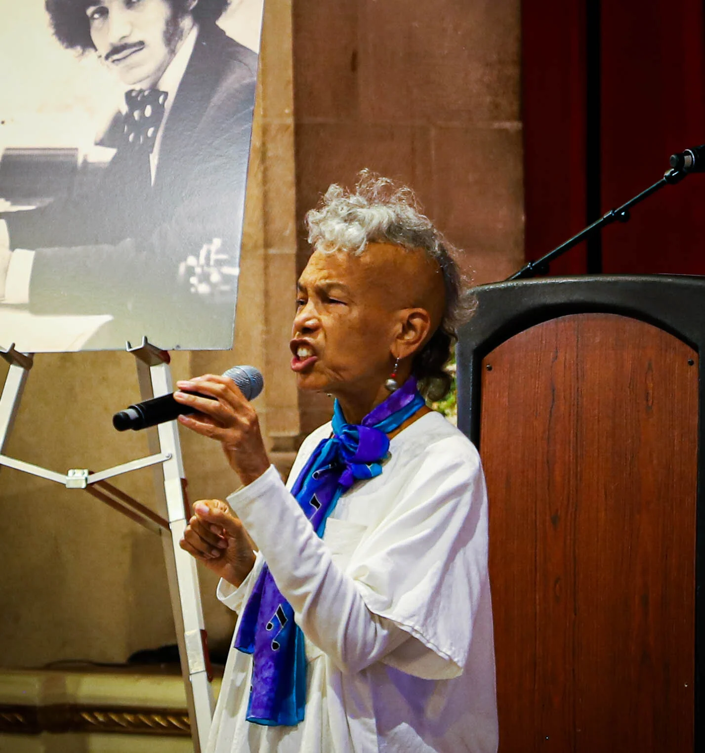 An elderly woman with a unique hairstyle holding a microphone and speaking at an event, with a black-and-white portrait of a man displayed on an easel beside her.