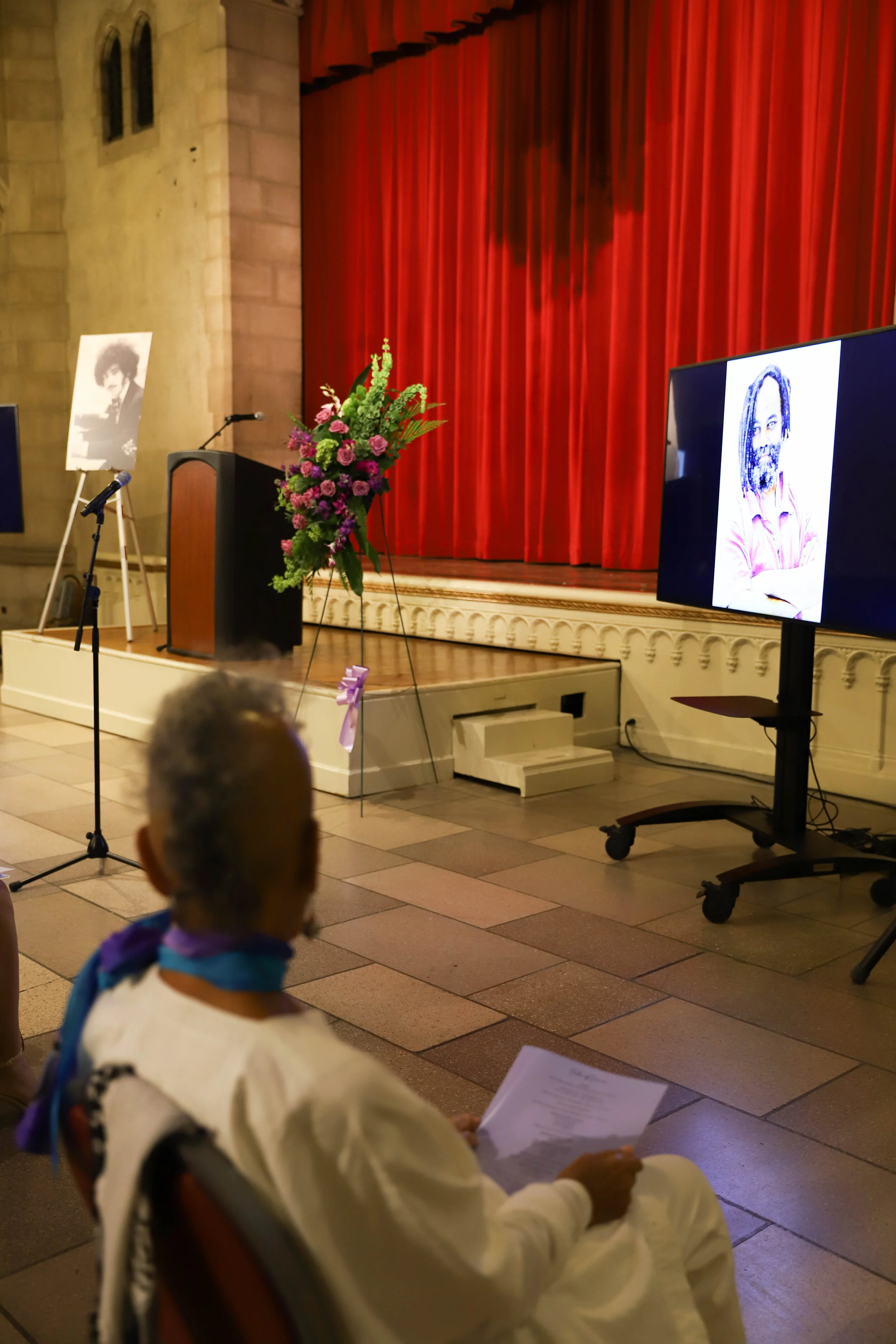 A woman dressed in a white traditional outfit is sitting and reading a paper while attending an event. The setting is a hall with a stage decorated with a large floral arrangement, a velvet red curtain backdrop, and two portraits displayed on screens