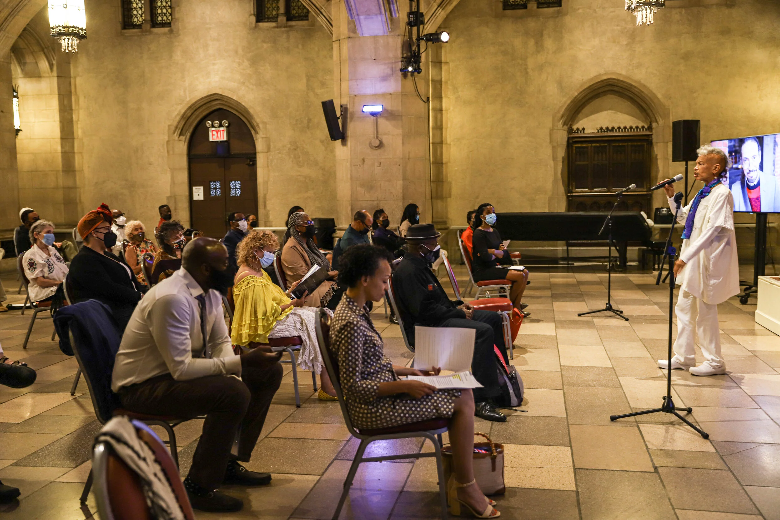 Group of people seated in an auditorium listening to a speaker holding a microphone. The speaker is a woman dressed in white, standing in front of a large screen displaying her image. The audience, wearing masks, is attentively watching the presentat