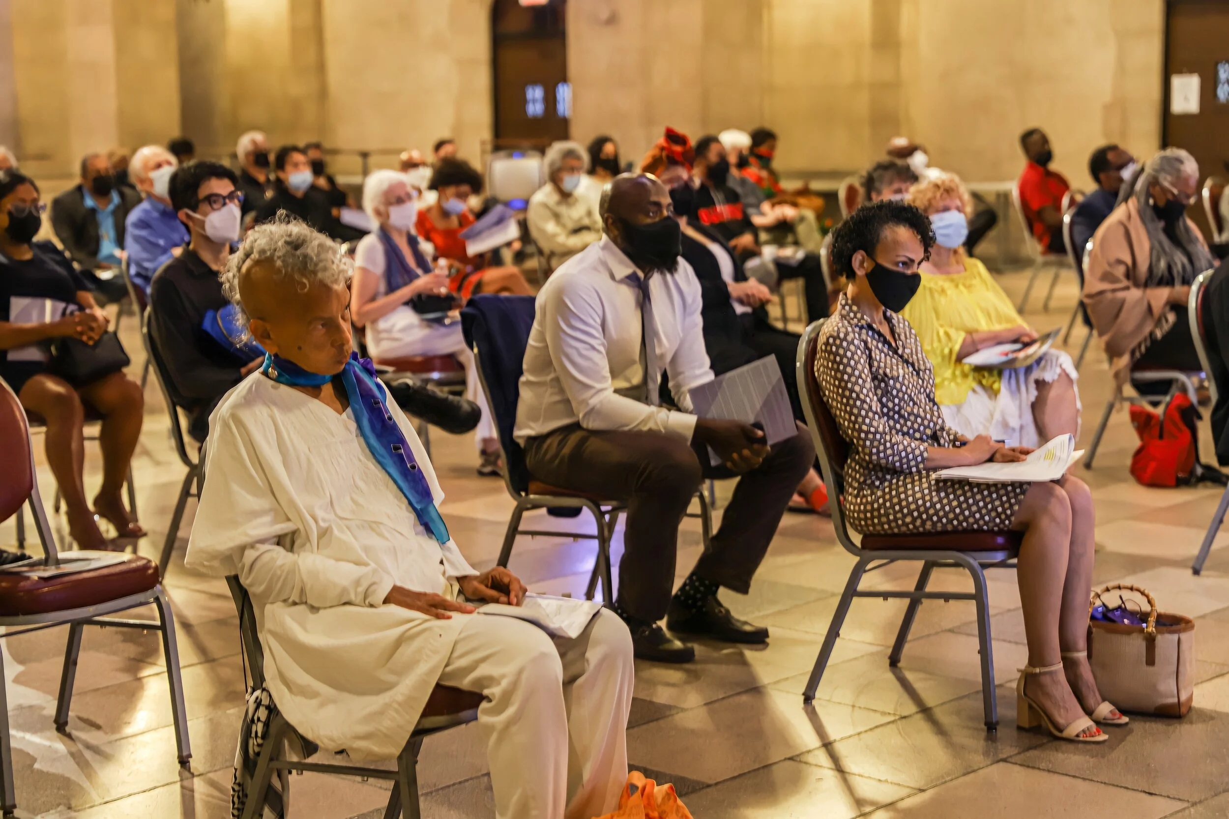 A diverse group of people sitting in chairs, attending an indoor event, all wearing masks, with some holding papers or notebooks.