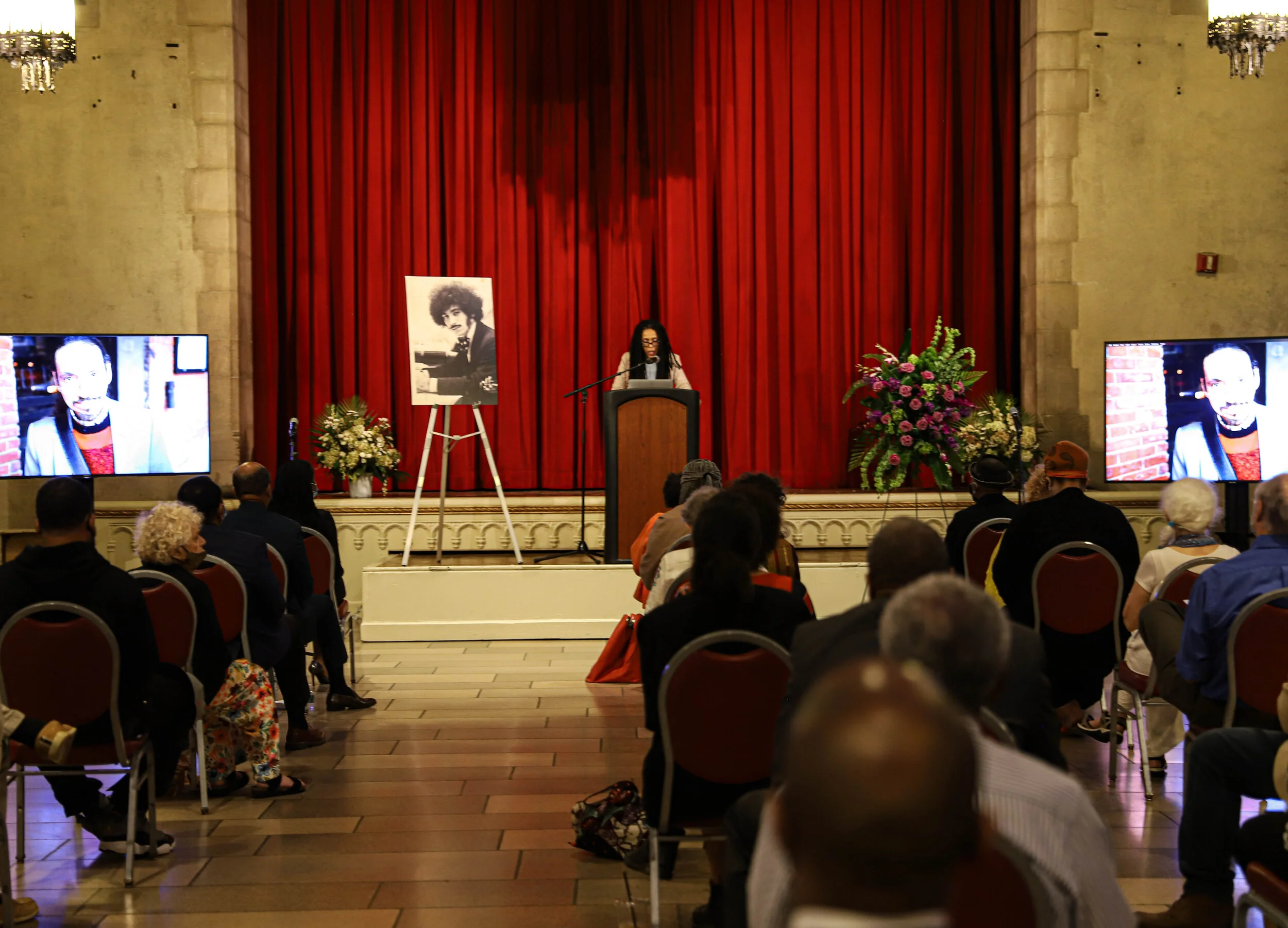 A woman is speaking at a podium on a stage with red curtains, flanked by floral arrangements and large photographs of a woman playing piano and a woman speaking in a television screen, with an audience seated in front.