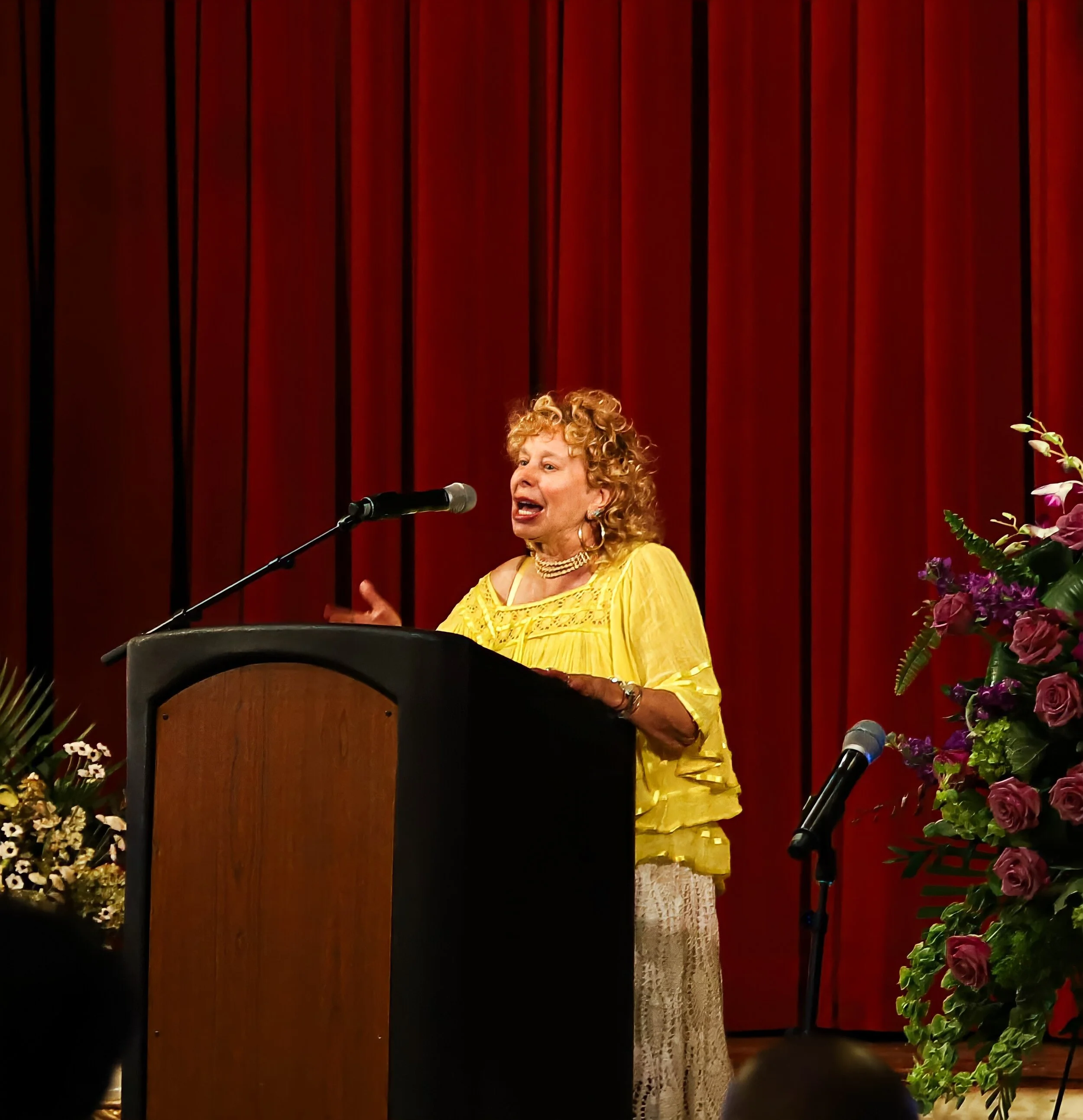 A woman in a yellow dress speaking at a podium with a microphone, surrounded by flowers, against a red curtain background.