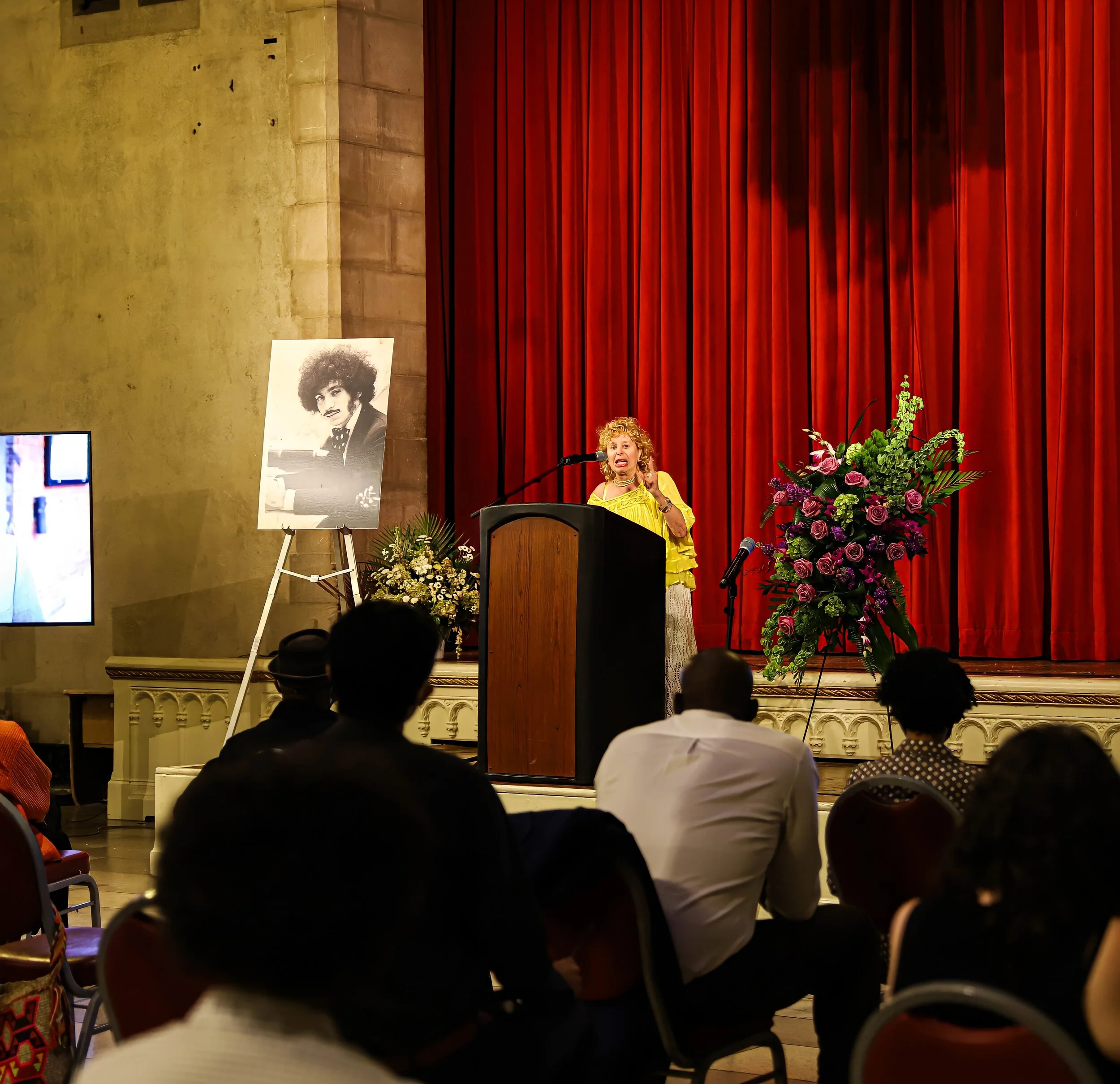 Woman speaking at a podium with a large floral arrangement in front, red curtains behind her, and an art photograph of a man's face on an easel to her left.