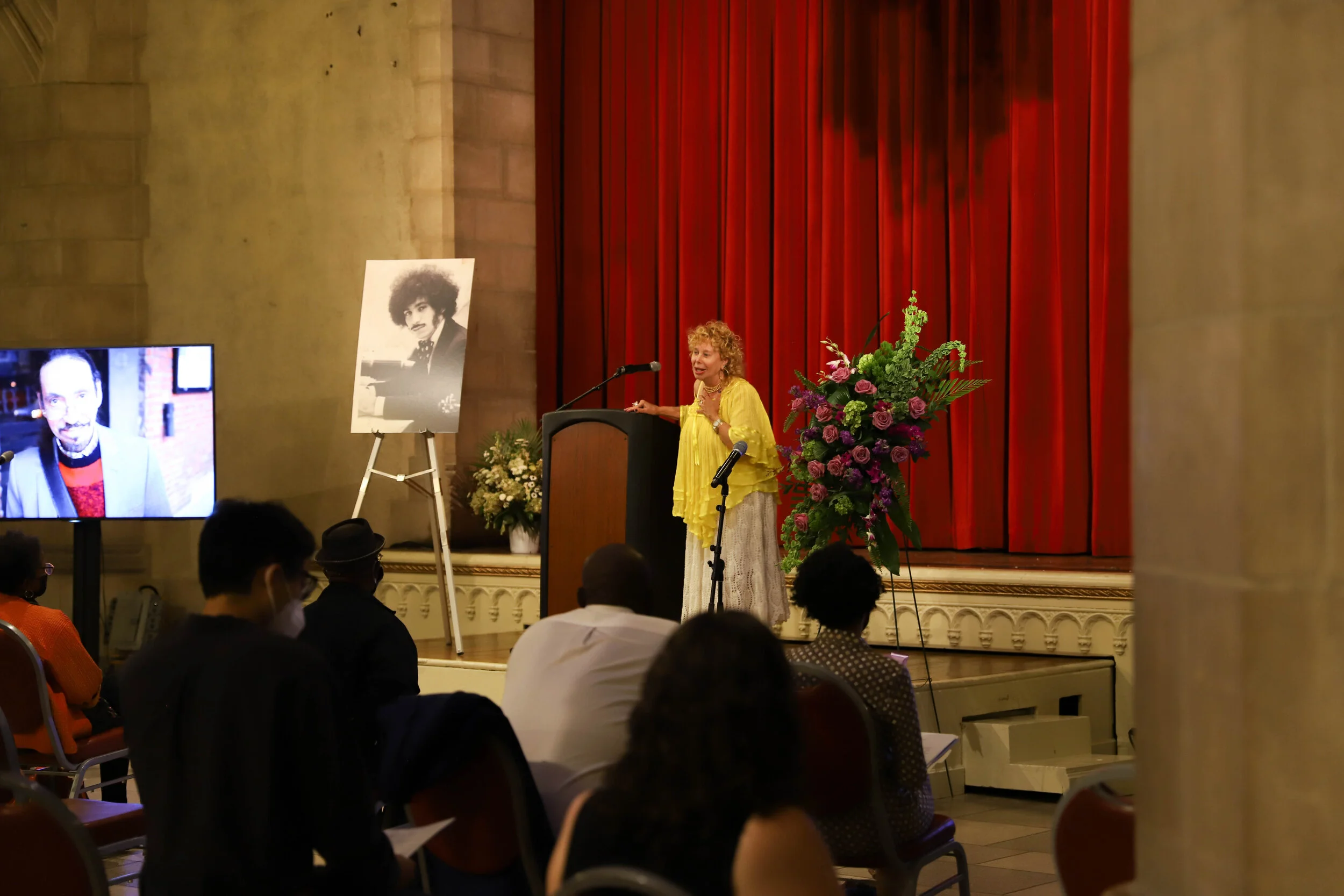 A woman with curly blonde hair wearing a yellow jacket giving a speech at a podium during an event, with a large floral arrangement beside her and an audience seated facing her. There are red curtains behind her, and a portrait of a man with curly ha