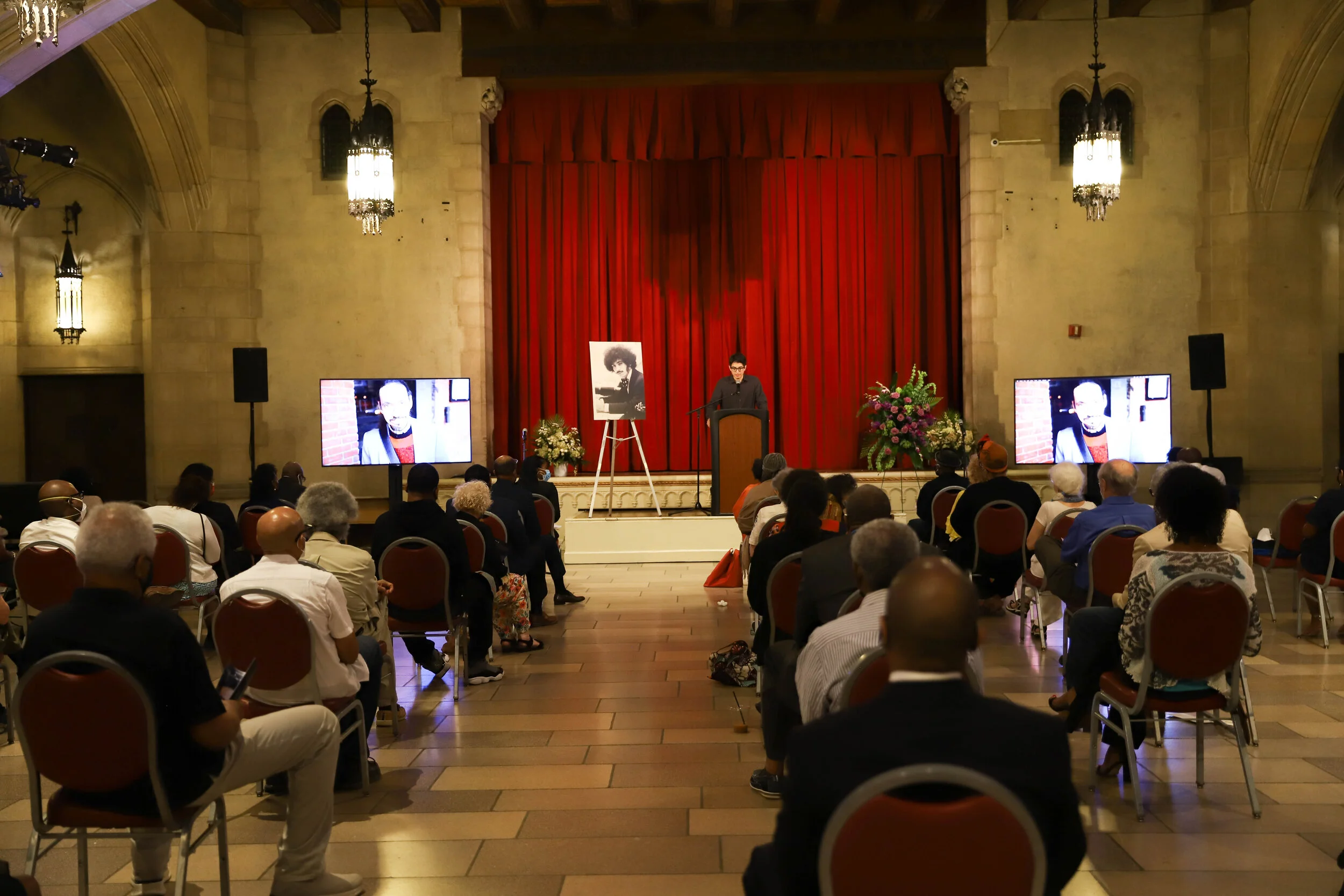 An event hall with a stage featuring a red curtain, people seated in chairs, multiple large screens displaying images of a person, and flowers on stage, with some attendees wearing masks.