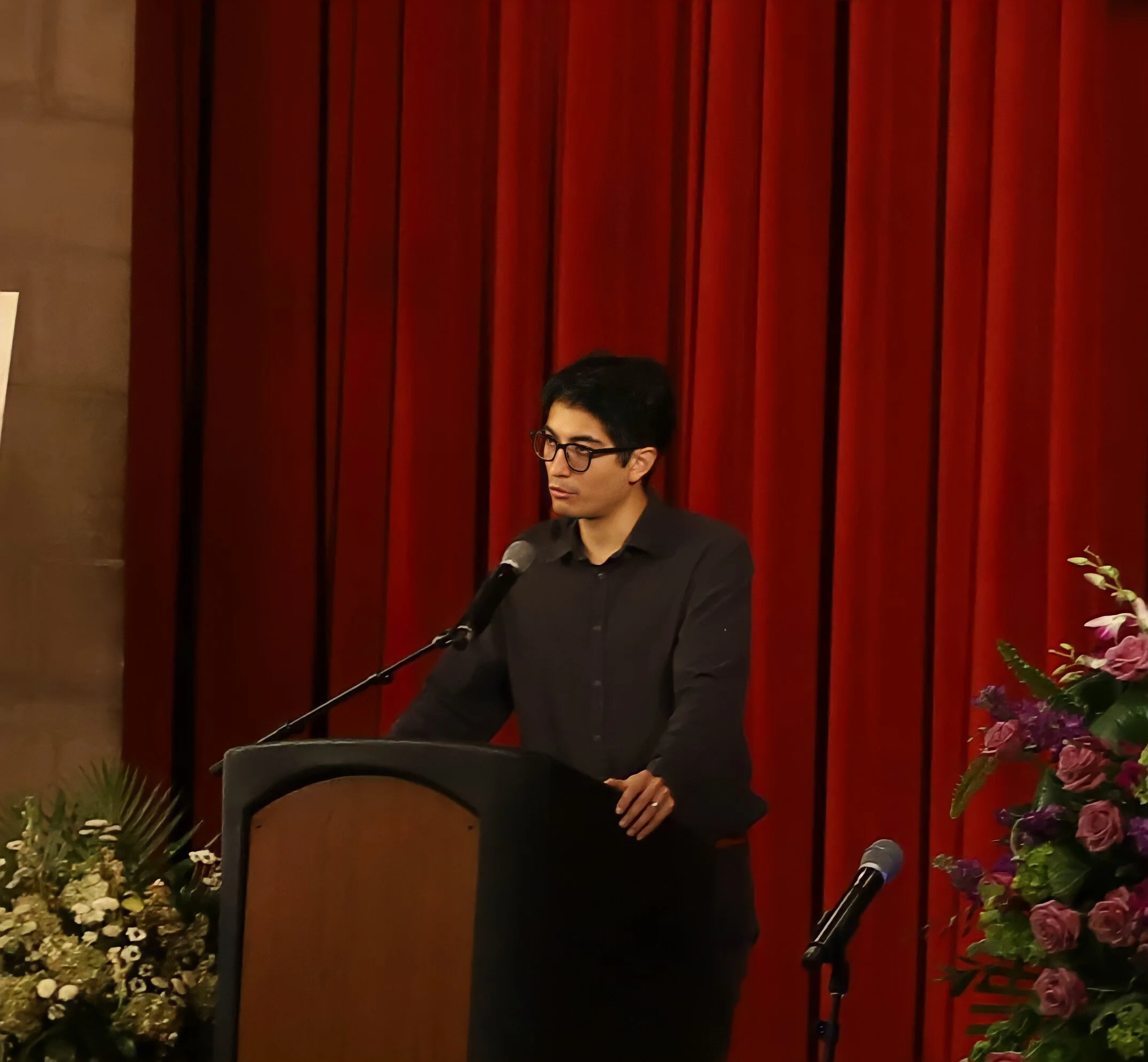 A person with short dark hair, glasses, and wearing a black shirt is standing at a podium in front of red curtains, speaking into a microphone. There are flowers on a table to their right.