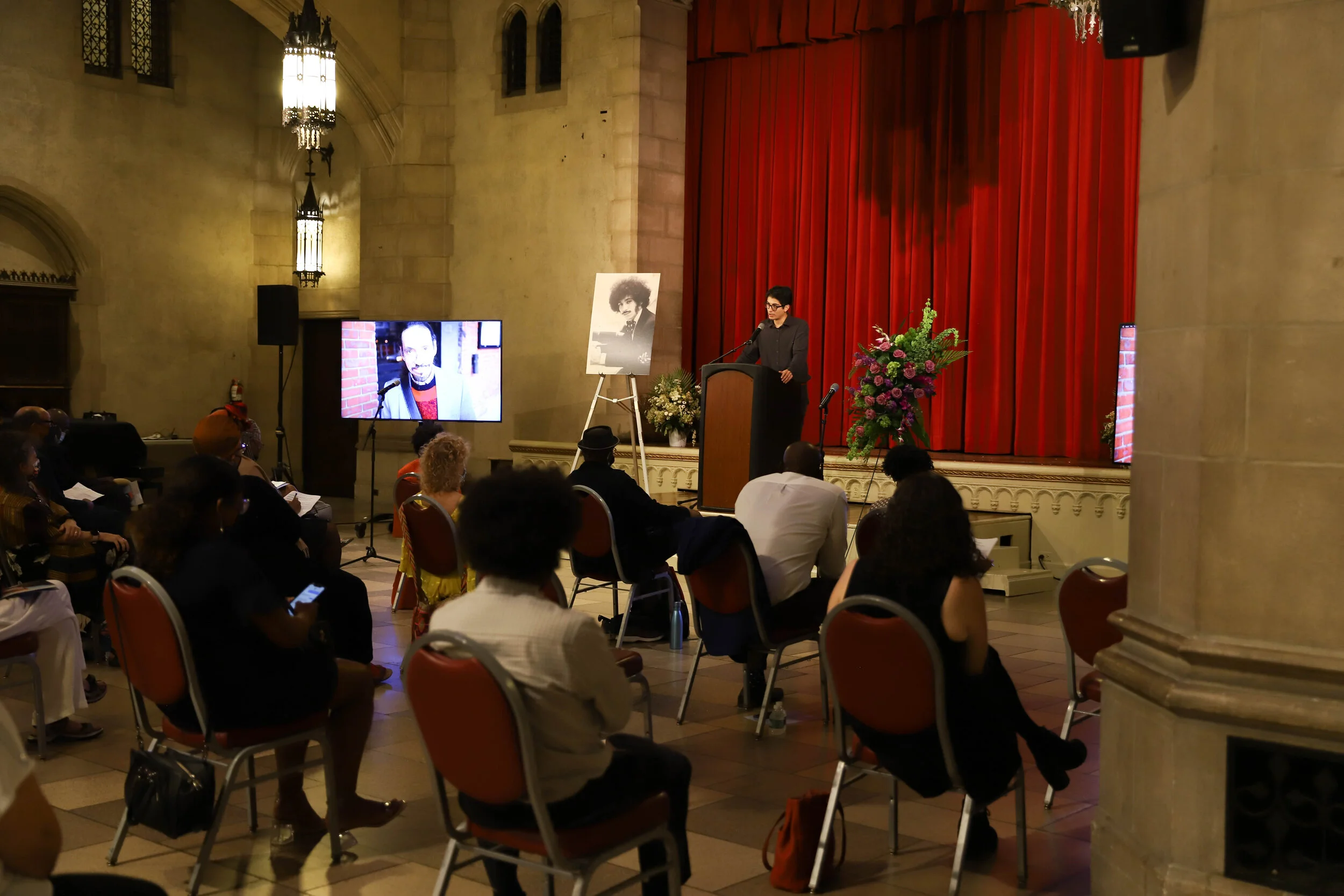 A woman in glasses and a dark gray blazer speaking at a wooden podium during a formal event in a large hall. Behind her, there is a large red curtain, and to the left, a poster of a person with curly hair and a large-screen displaying a close-up of a