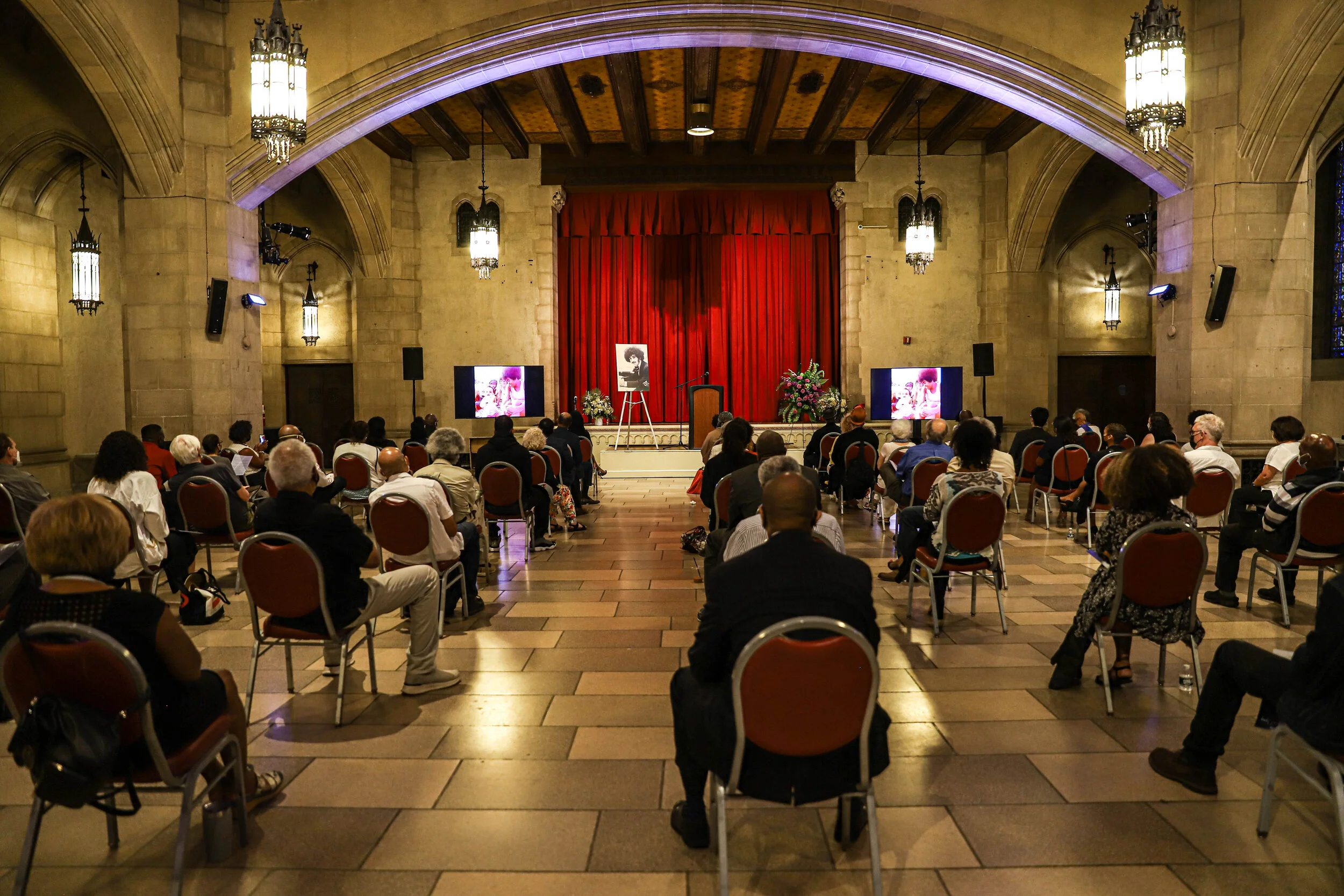 Indoor event space with an audience seated facing a stage with a red curtain, large floral arrangement, microphones, and two screens displaying images, set in a historic hall with stone walls and ornate hanging lights.