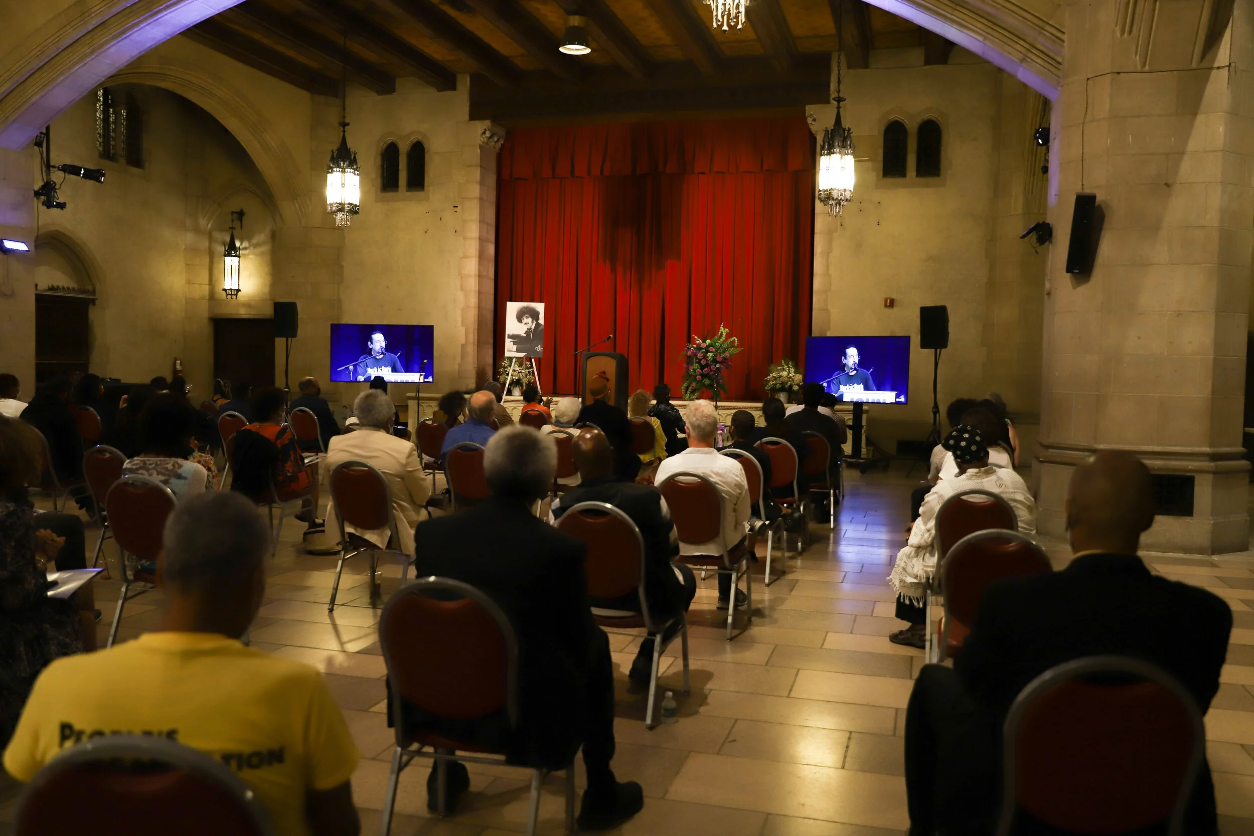 Interior of a large hall with stone walls and a wooden vaulted ceiling, decorated with chandeliers and wall sconces. The hall has an elevated stage with red curtains and two large screens on either side showing a person speaking. There are chairs fil