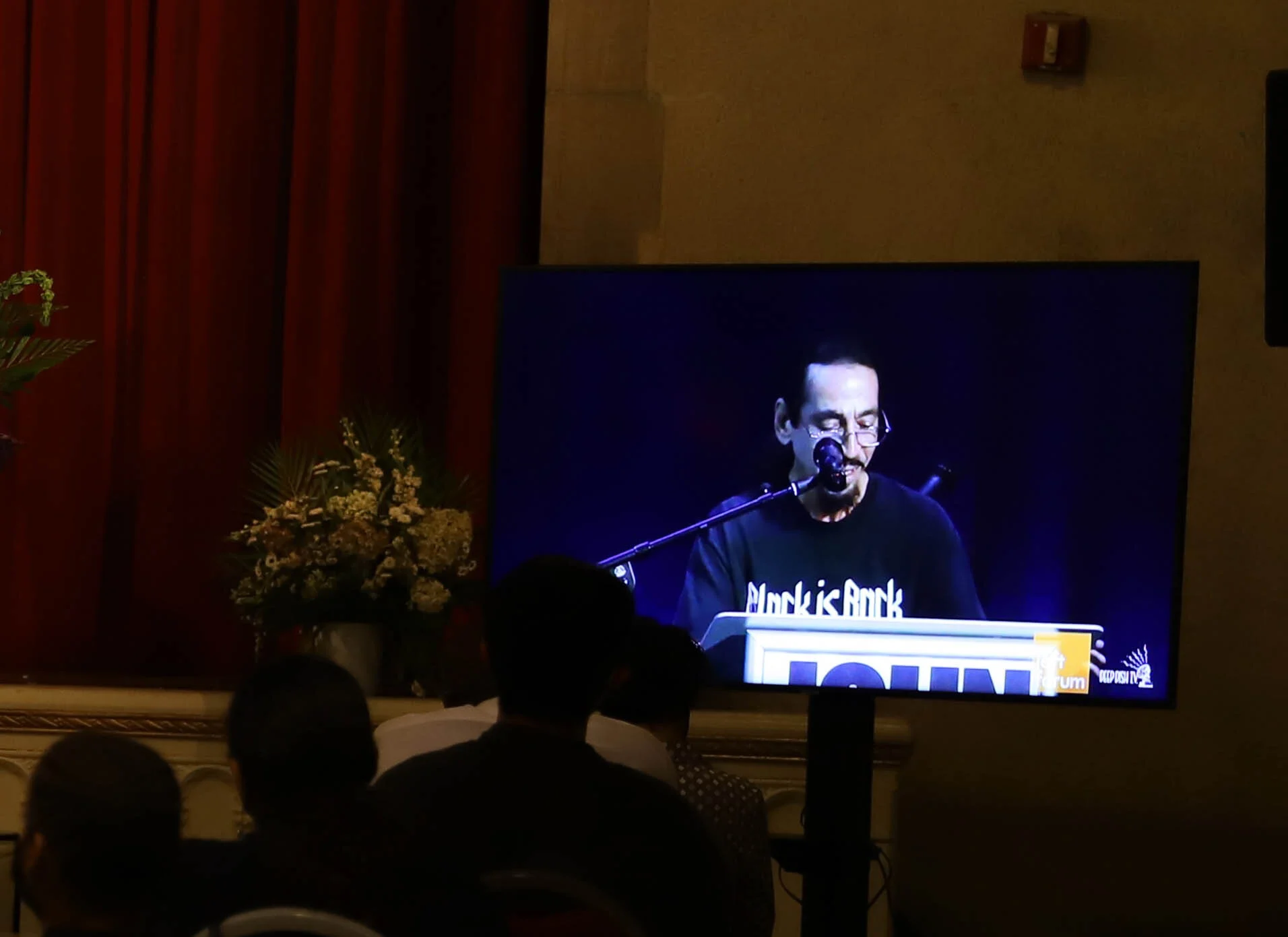 A man with glasses and dark hair playing a piano on stage at an event, wearing a black t-shirt with white text. The stage has red curtains, flowers in vases on either side, and a large screen displaying the man. The audience is visible in silhouette.