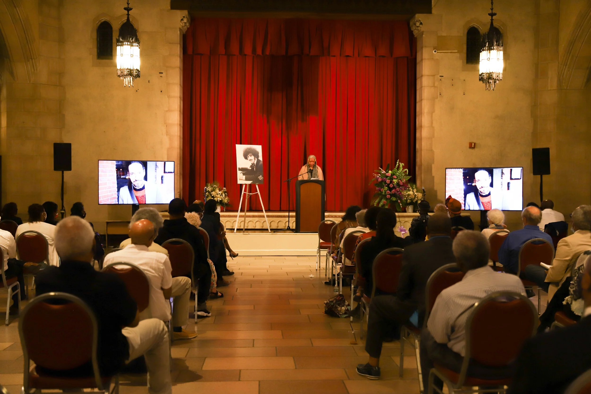 A woman giving a speech at a podium during a formal event in a large hall with red curtains. Large floral arrangements and two large screens displaying images are on either side of the stage. Audience members are seated facing the stage.