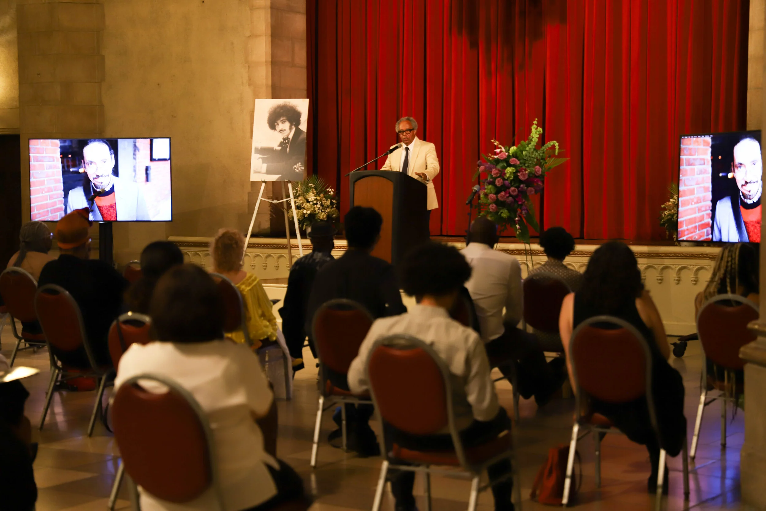 A man in a beige jacket and black tie standing at a podium, speaking to an audience at a formal event. There are two large screens displaying a photo of a young man with curly hair and a mustache. A framed black-and-white picture of the same young ma