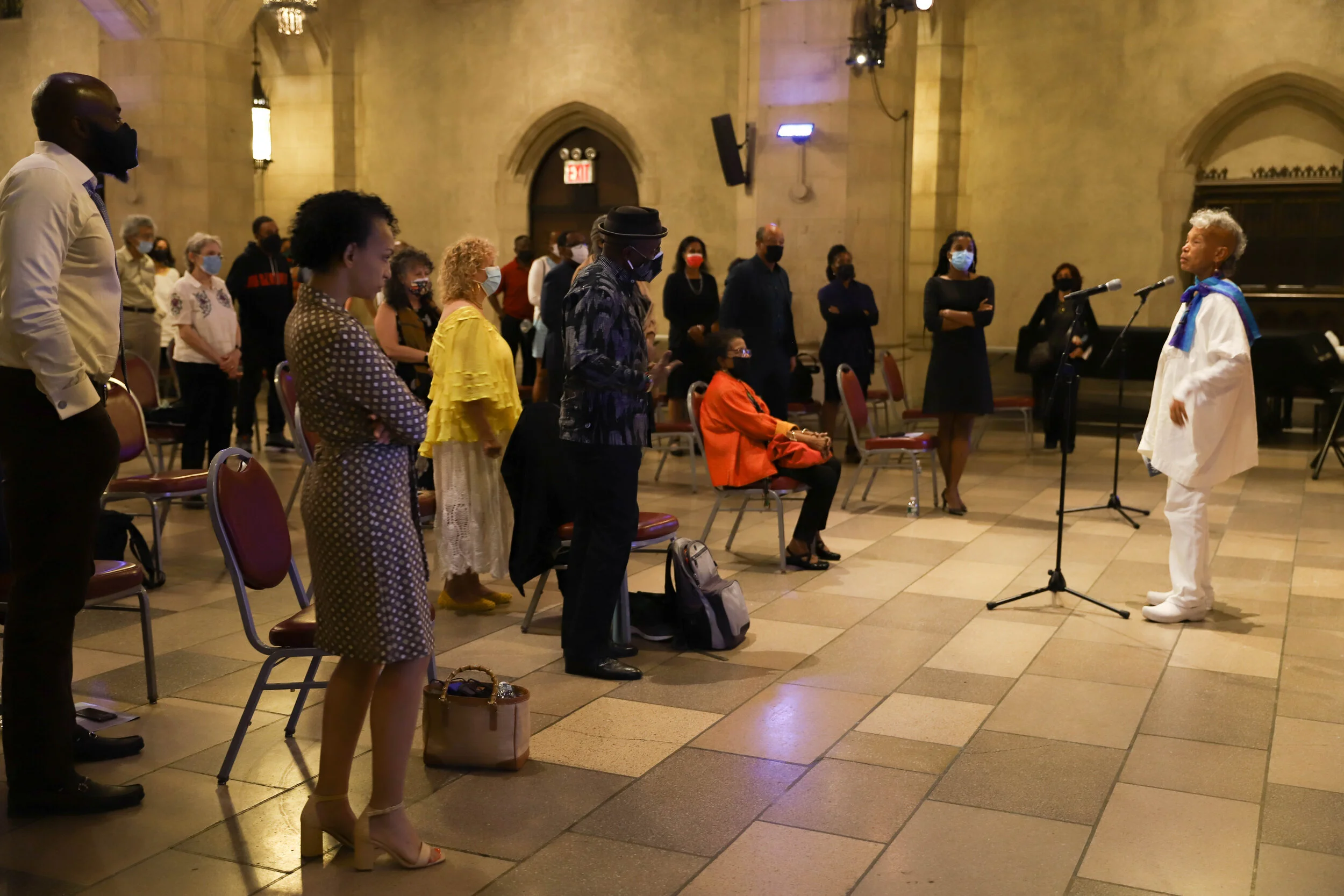 An indoor event with people attending a. standing in a hall with high arched ceilings. Several individuals are standing or sitting, and most are wearing face masks. A person dressed in a white academic gown with a blue scarf stands at a microphone, p