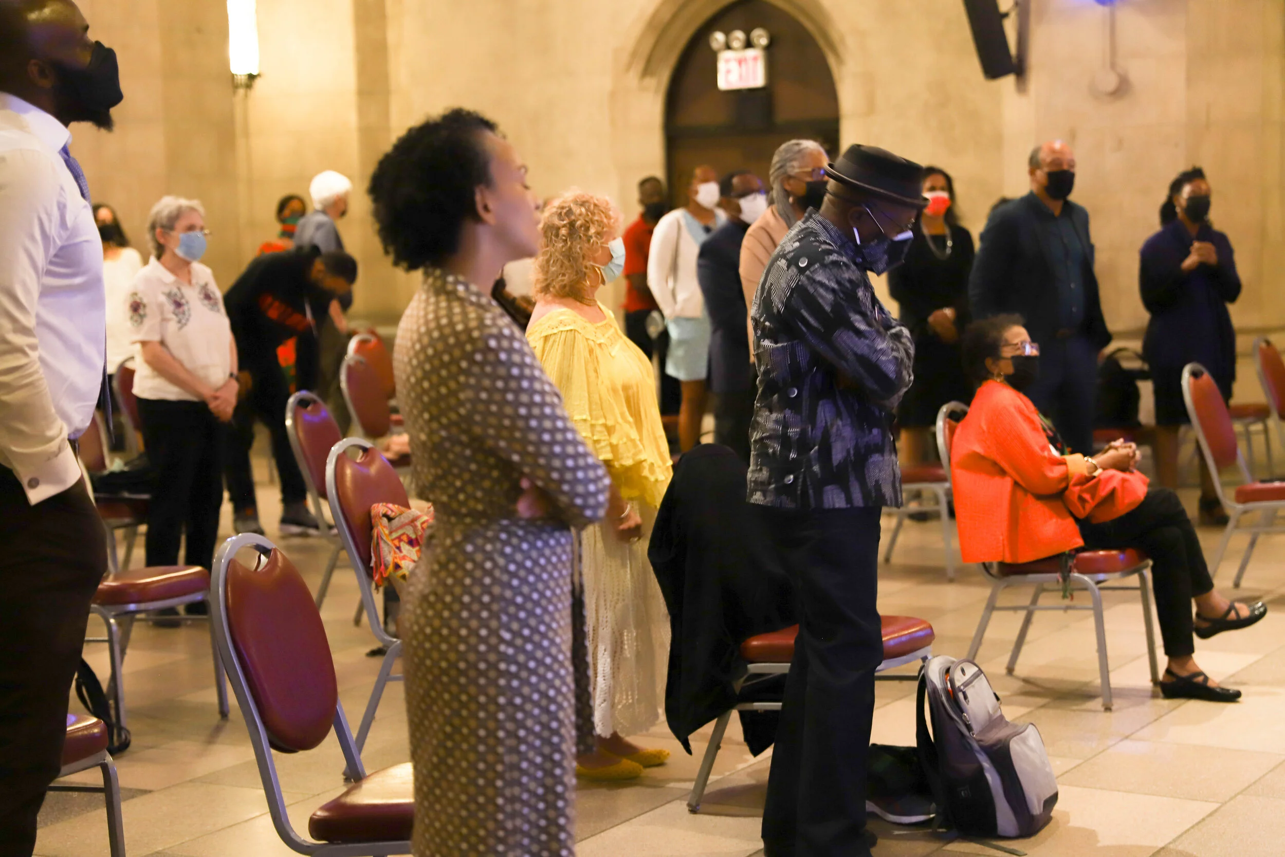 People attending a prayer or memorial service in a church, standing with heads bowed and eyes closed, wearing face masks.