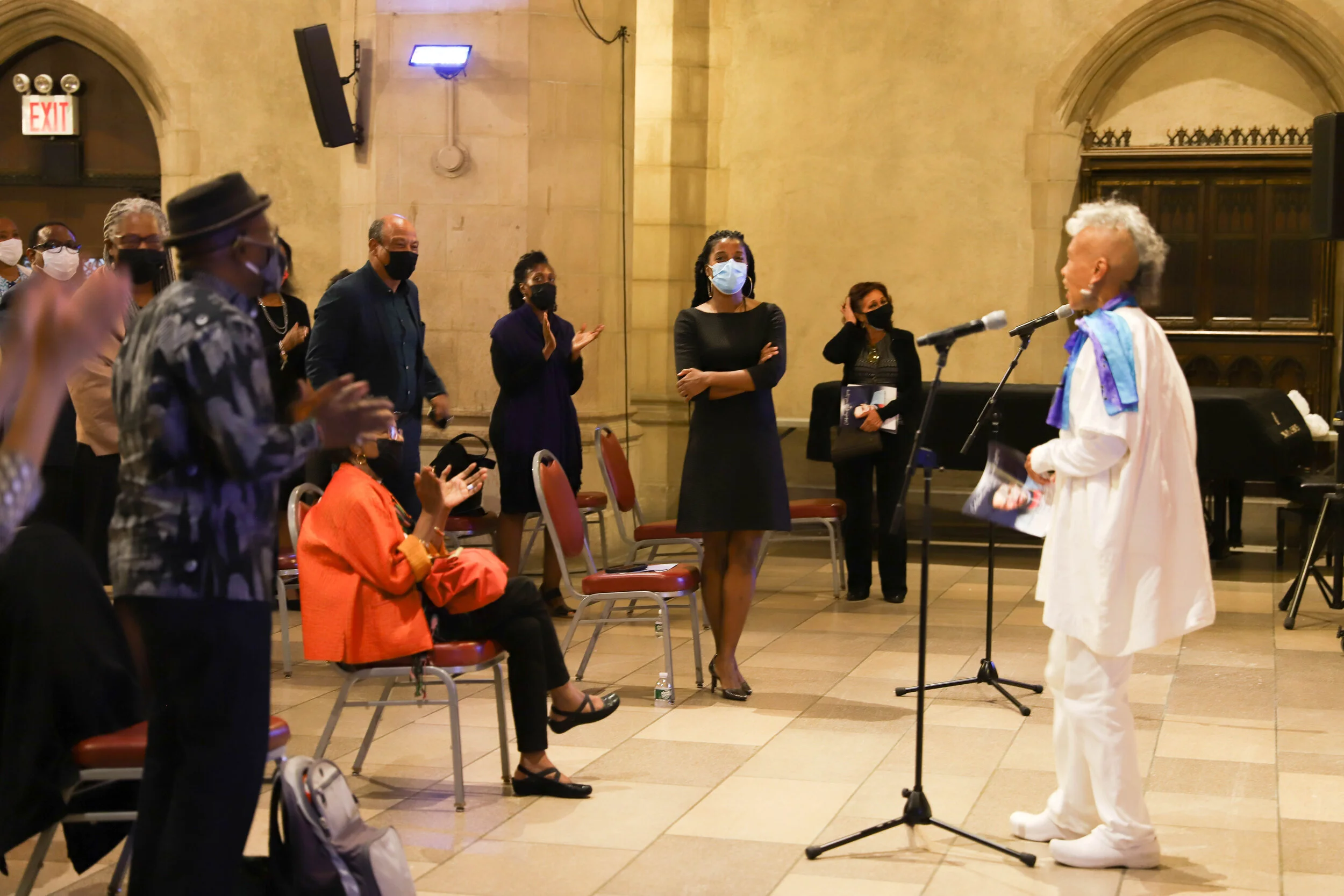 A diverse group of people wearing masks is gathered in a historic building with stone walls and arched doorways. They are listening to a speaker, an older woman with curly gray hair, dressed in white, standing in front of a microphone.