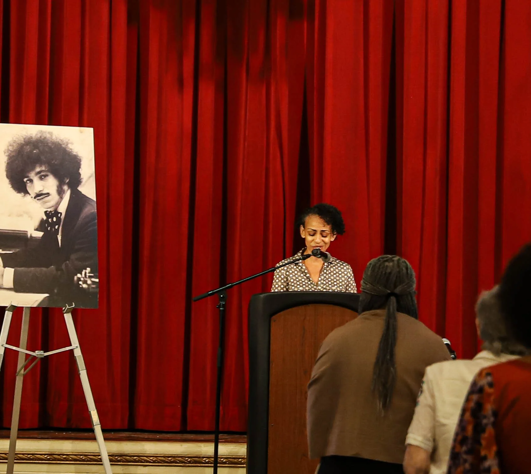 A woman singing at a microphone on stage with a red curtain backdrop, with an audience watching, and a black-and-white photo of a man playing piano displayed on an easel to her left.