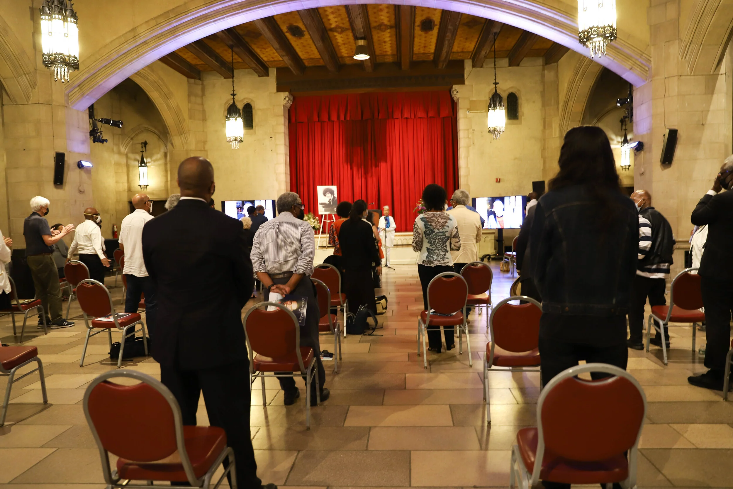 A gathering of people in a large hall with stone walls, arched ceilings, and ornate hanging lamps. Some attendees are standing, wearing masks, and facing a stage with a red curtain, where a speaker is present with portraits and floral arrangements.