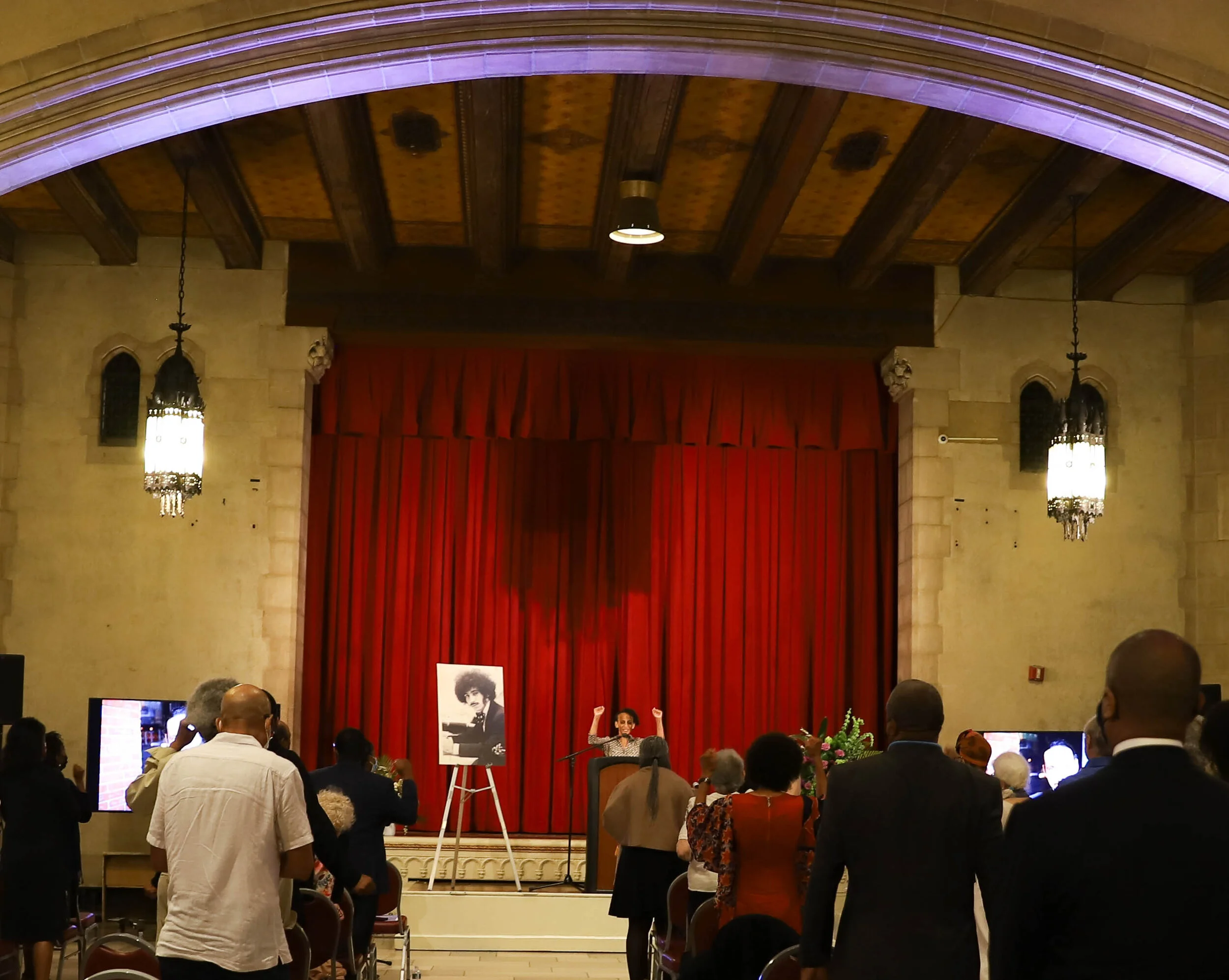 Inside a concert hall with a stage decorated with red curtains, a woman is speaking at a microphone. People are gathered, some standing and some seated, listening to her. There is an easel with a large black-and-white portrait of a young man with an 
