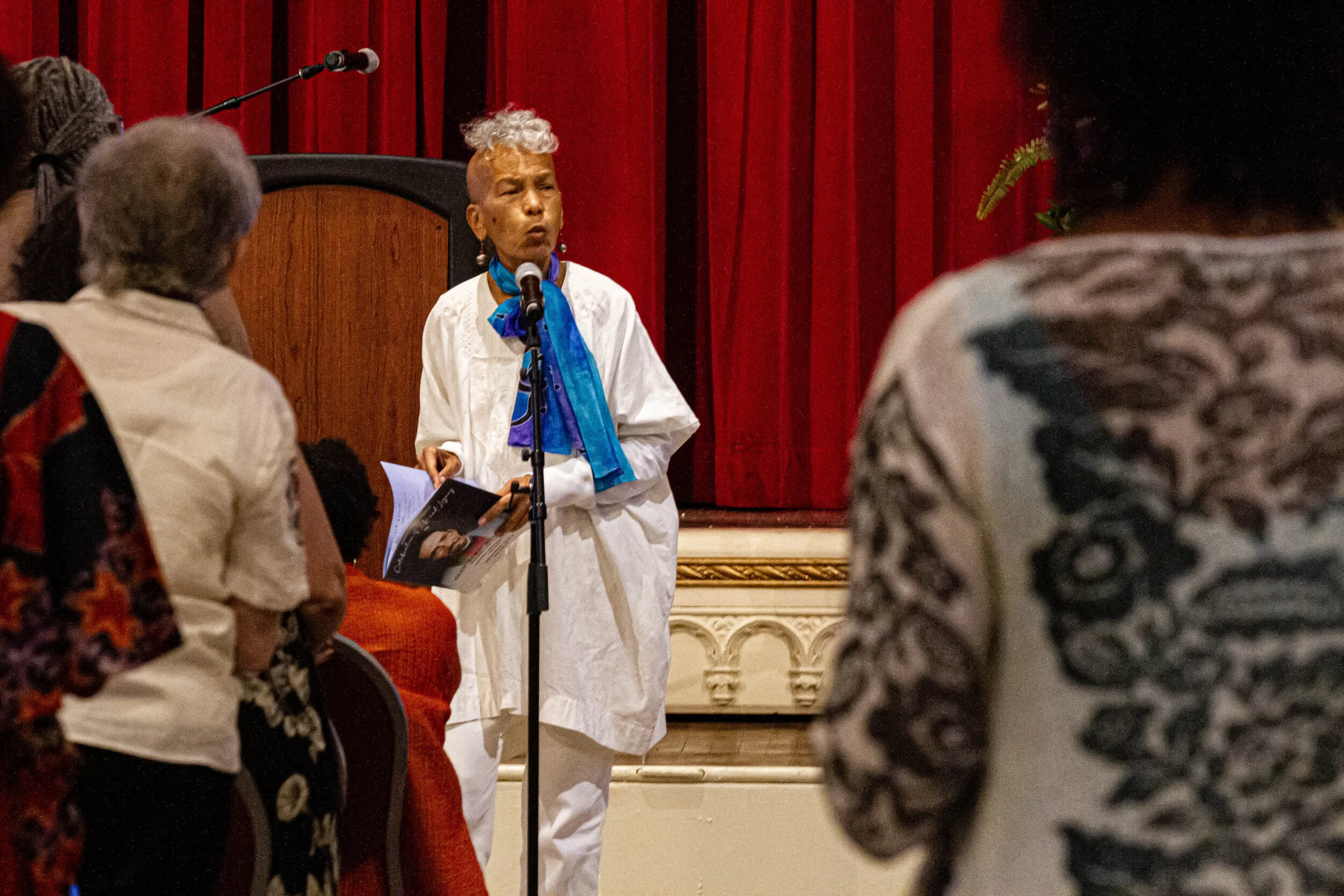 A woman with gray and curly hair, wearing white traditional attire and a blue scarf, is speaking at a microphone in front of an audience. The setting appears to be an indoor stage with red curtains and decorative molding.