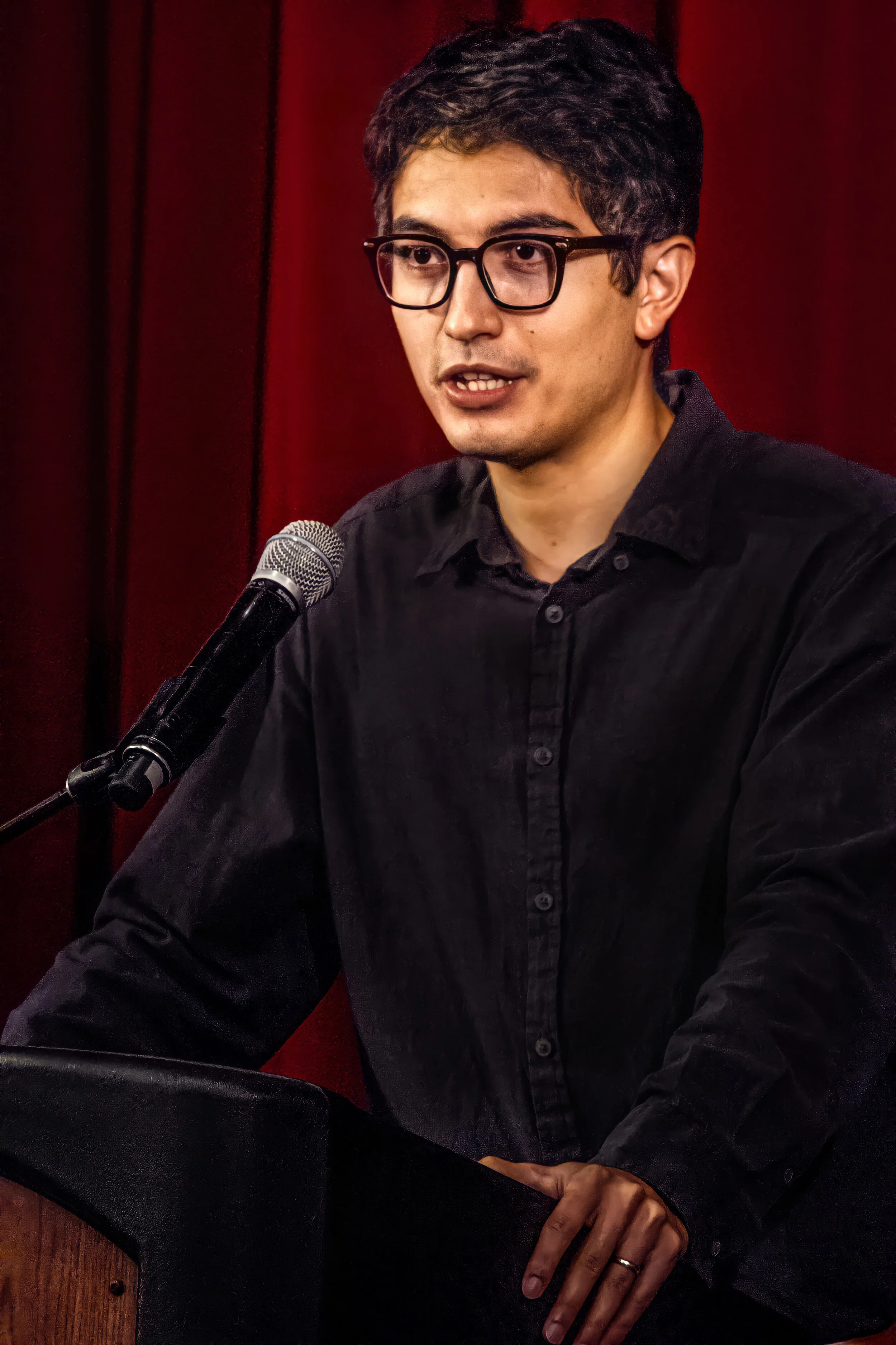 A young man with dark hair, glasses, and wearing a black shirt is speaking into a microphone during a recording or podcast session. He is seated at a table with a red curtain or wall in the background.