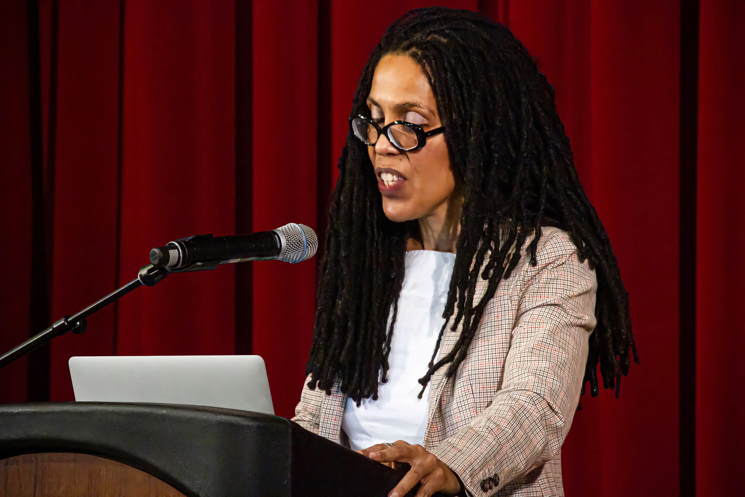 A woman with long dreadlocks and glasses speaking into a microphone at a podium in front of red curtains.