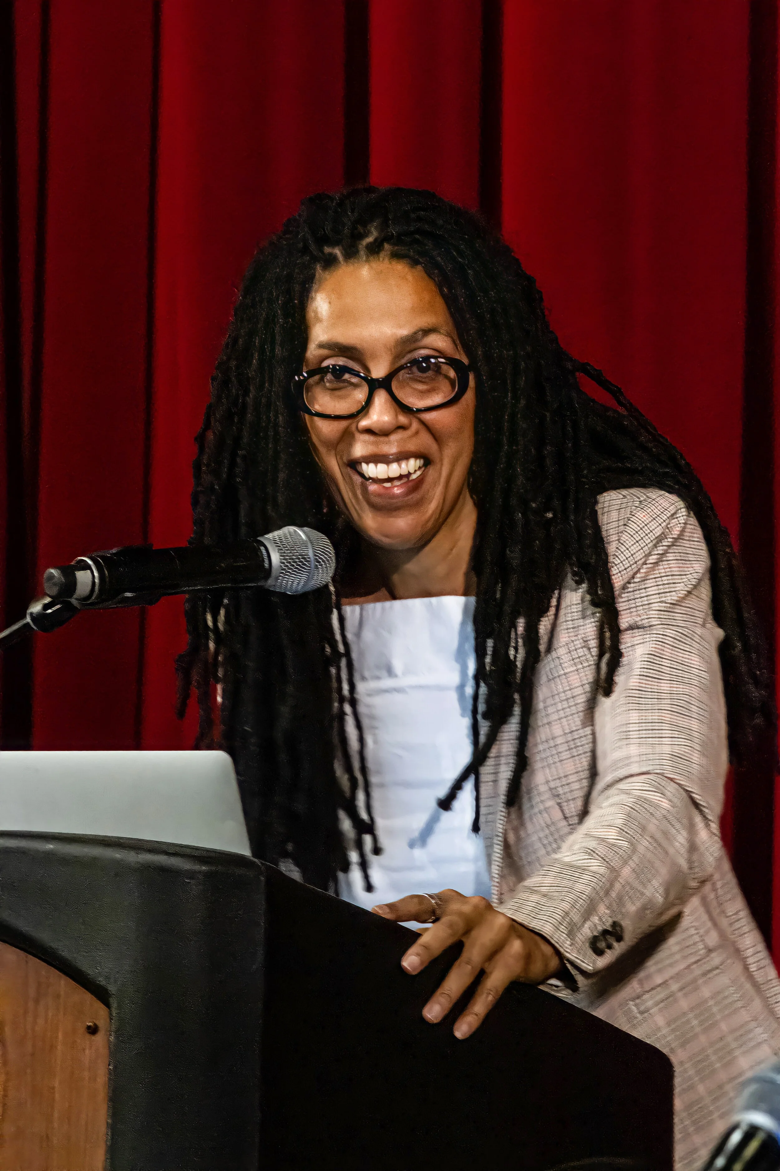 A woman with glasses, long dreadlocks, playing a keyboard and smiling, sitting in front of a red curtain.