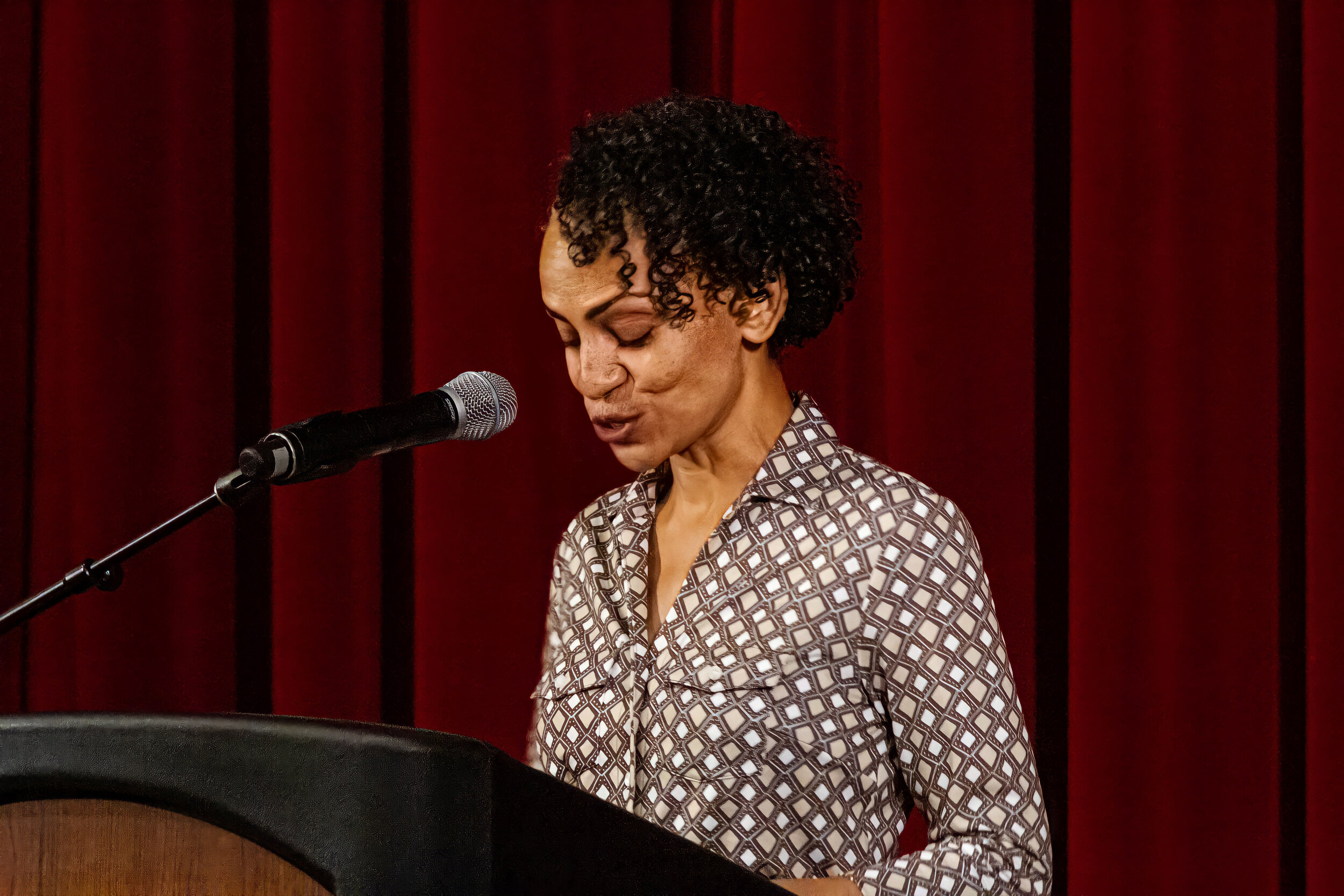 A woman with short curly black hair performing into a microphone on stage, wearing a patterned blouse, with a red curtain backdrop.