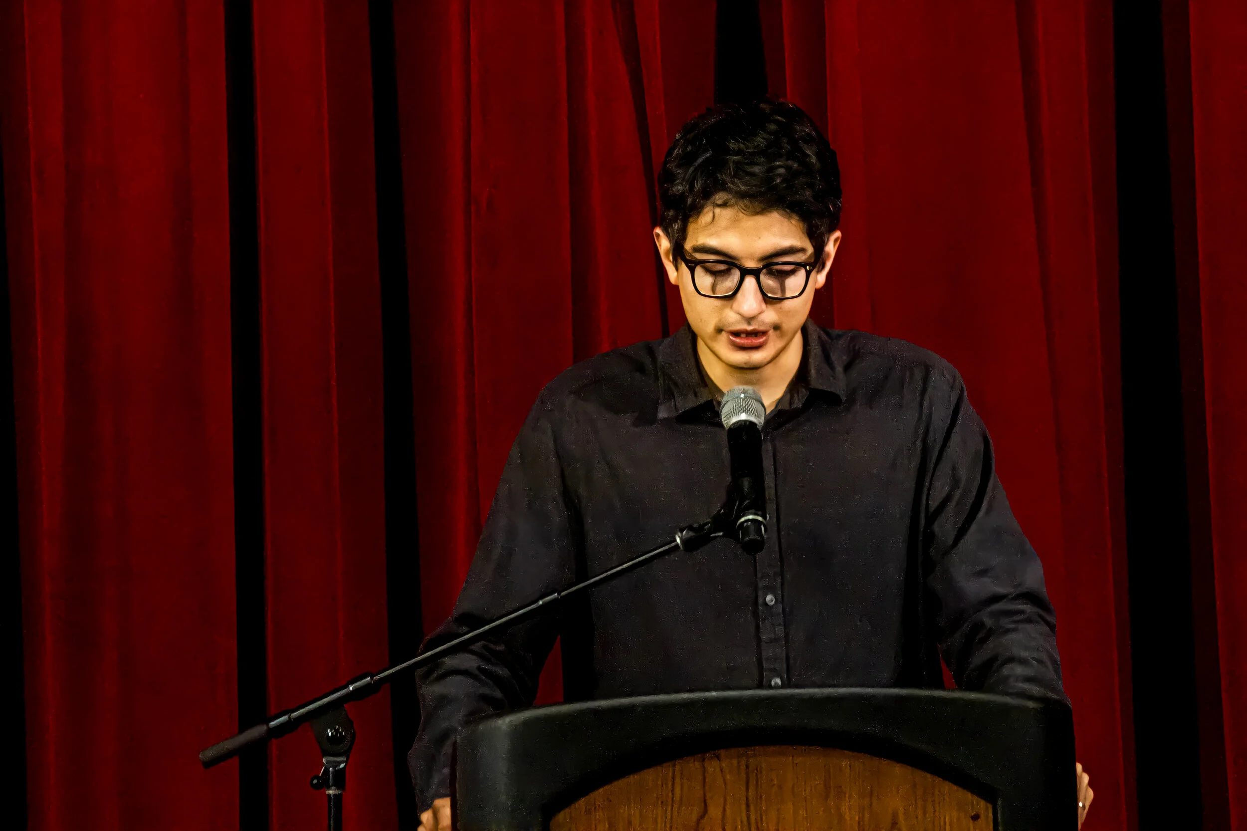 Young man with black curly hair and glasses speaking at a podium in front of red curtains.