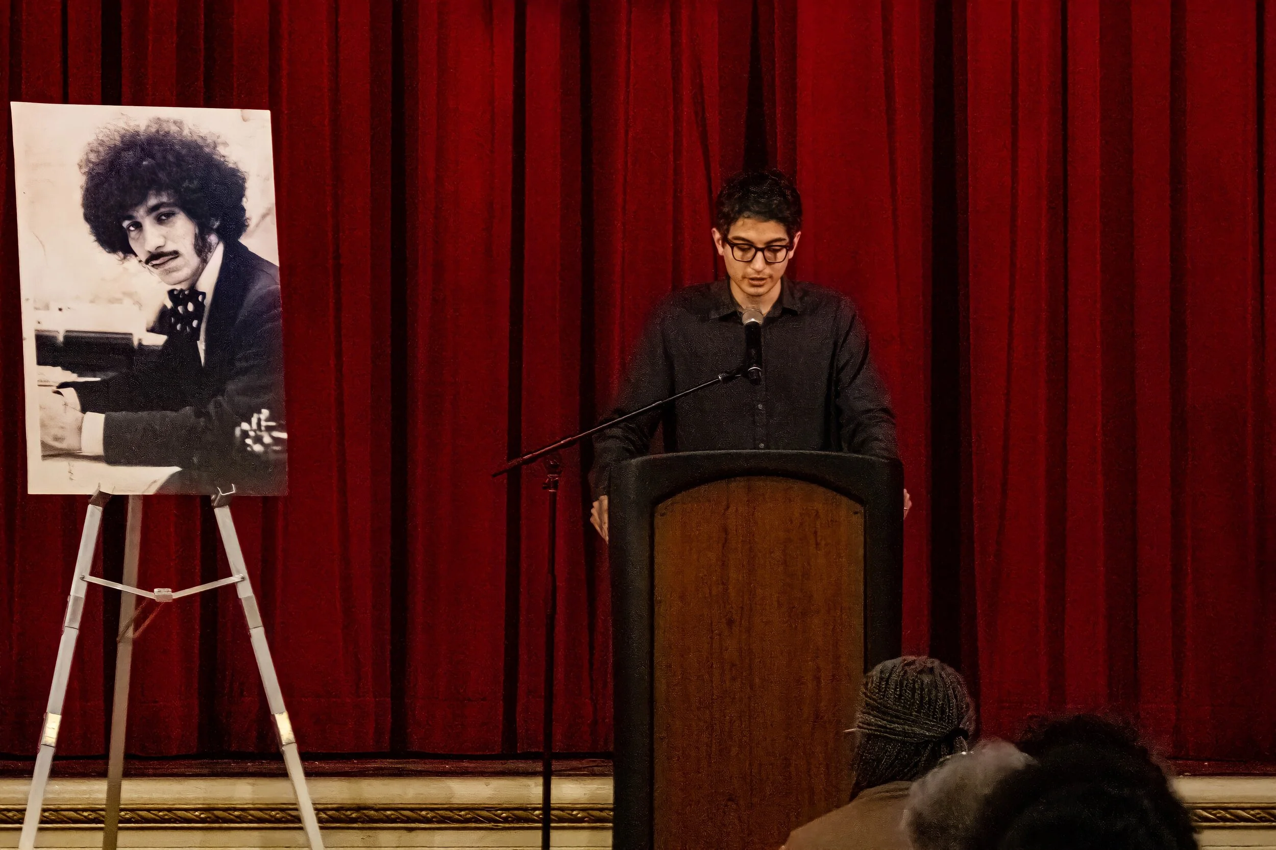 A young man wearing glasses and a black shirt stands at a podium with a microphone on stage in front of red velvet curtains. To his left, a black-and-white poster of a man with curly hair and a mustache playing the piano is displayed on an easel.