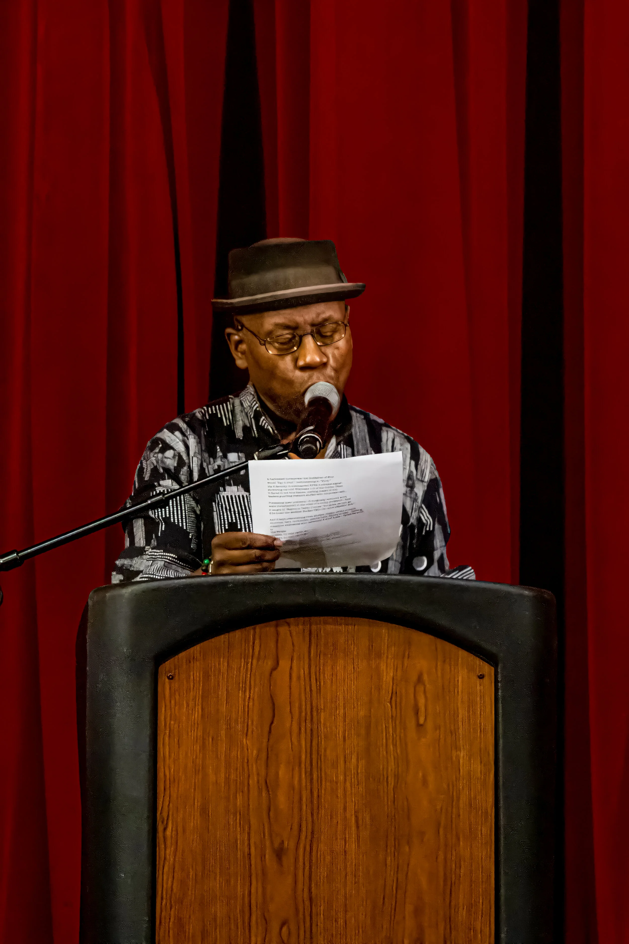 A man wearing glasses, a black hat, and a patterned shirt is standing at a podium reading from a piece of paper, in front of red curtains.