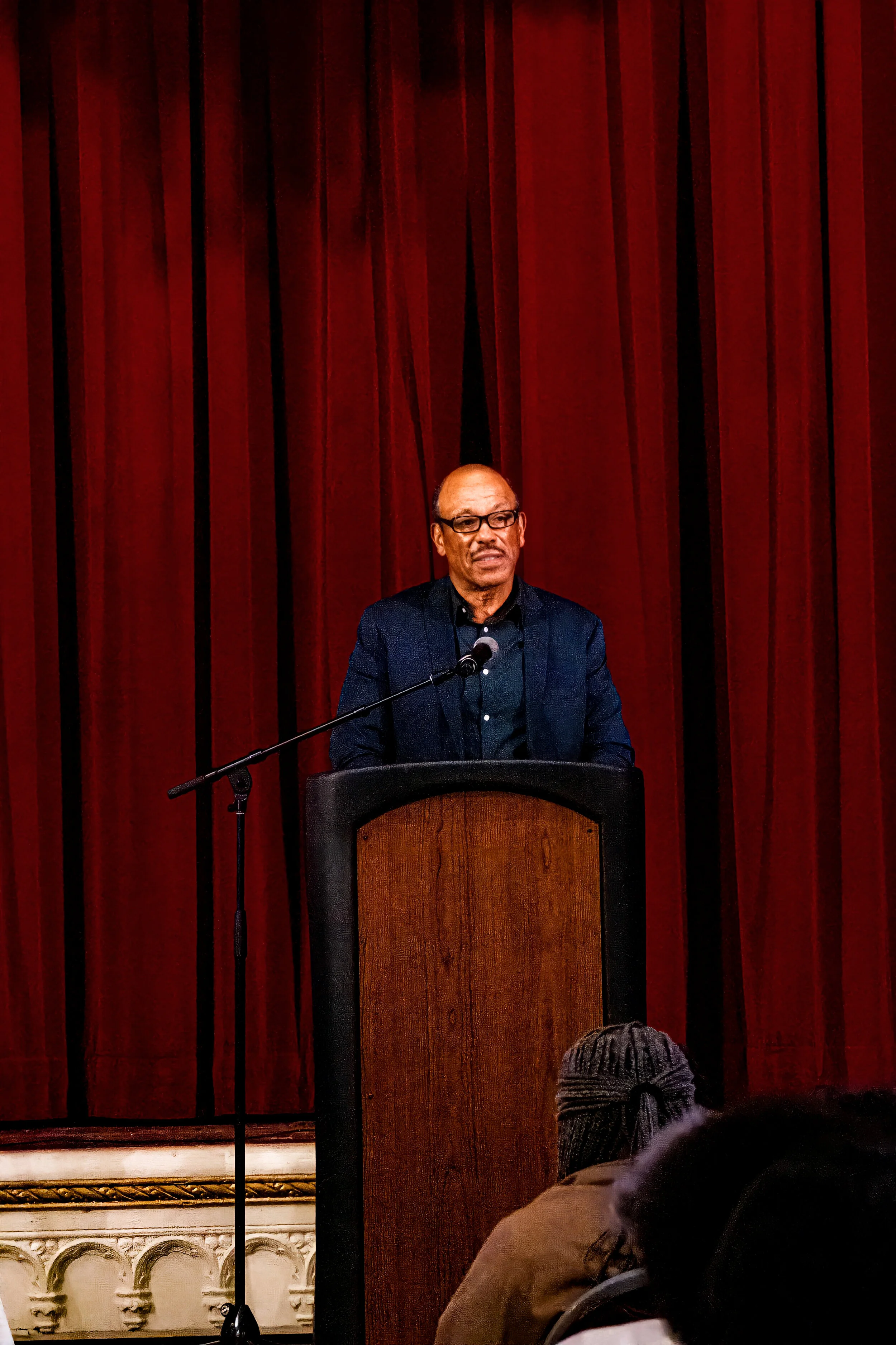 A man in glasses and a dark jacket stands behind a podium and speaks into a microphone, with a red curtain in the background and audience members in the foreground.