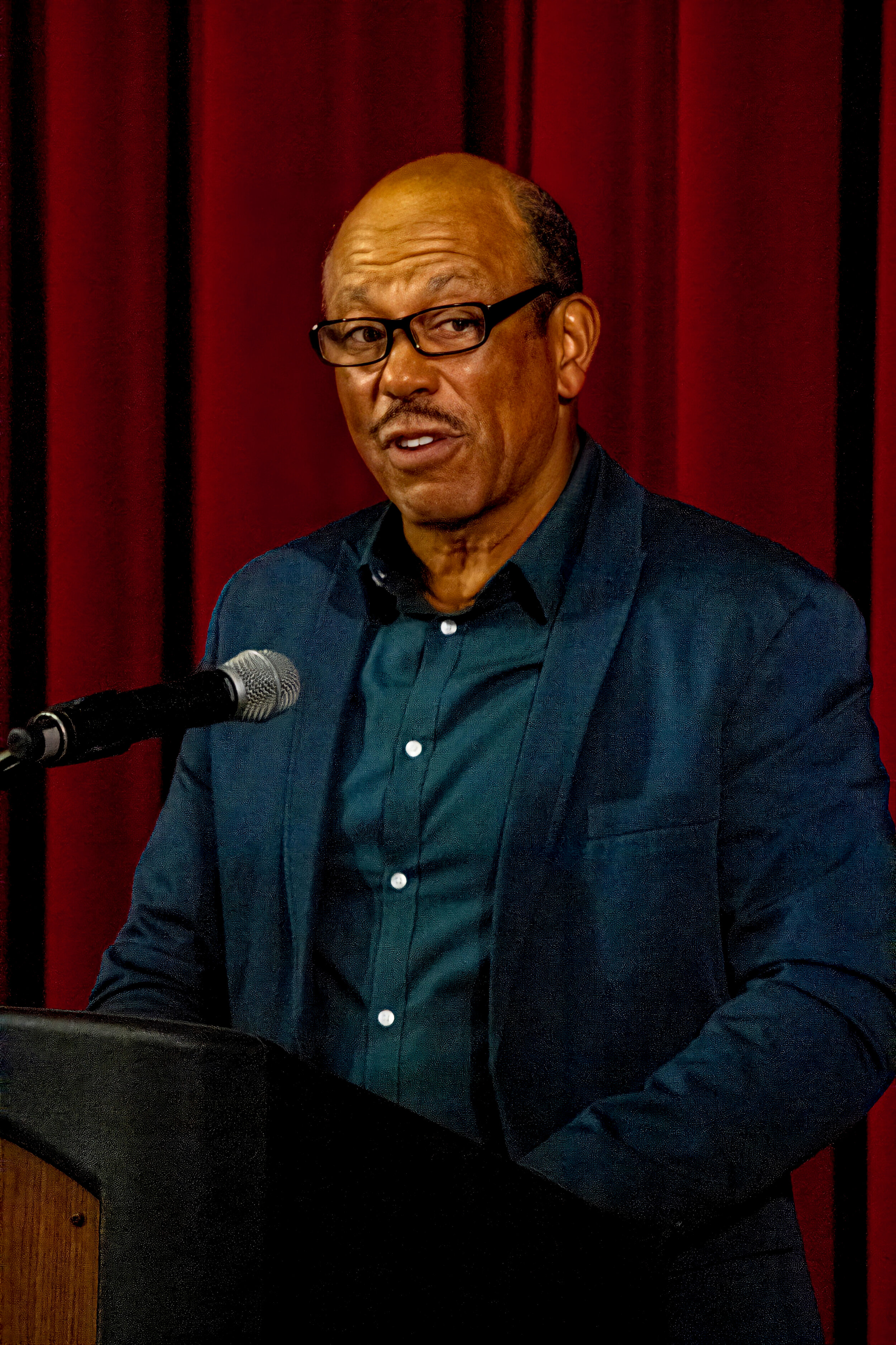 A middle-aged man wearing black-rimmed glasses and a dark blue suit is speaking into a microphone in front of a deep red curtain.