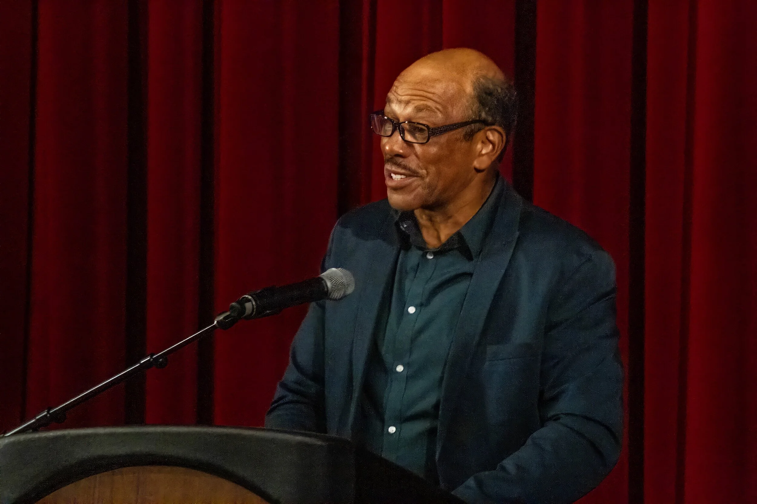 A man speaking or singing into a microphone with a red curtain backdrop.