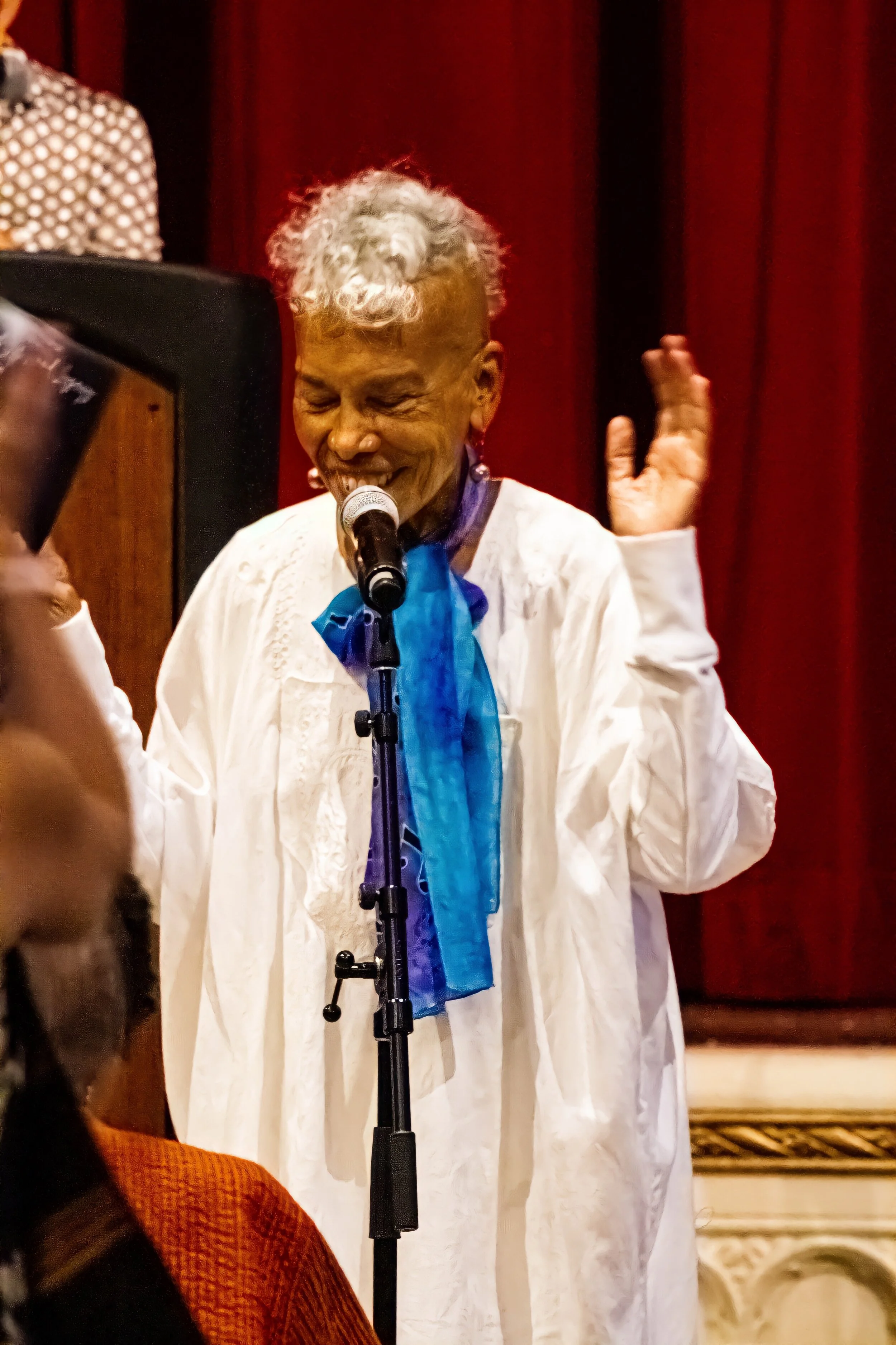 An elderly woman with curly blonde hair performing on stage, singing into a microphone and wearing a white dress with a blue scarf. Red curtains are in the background.