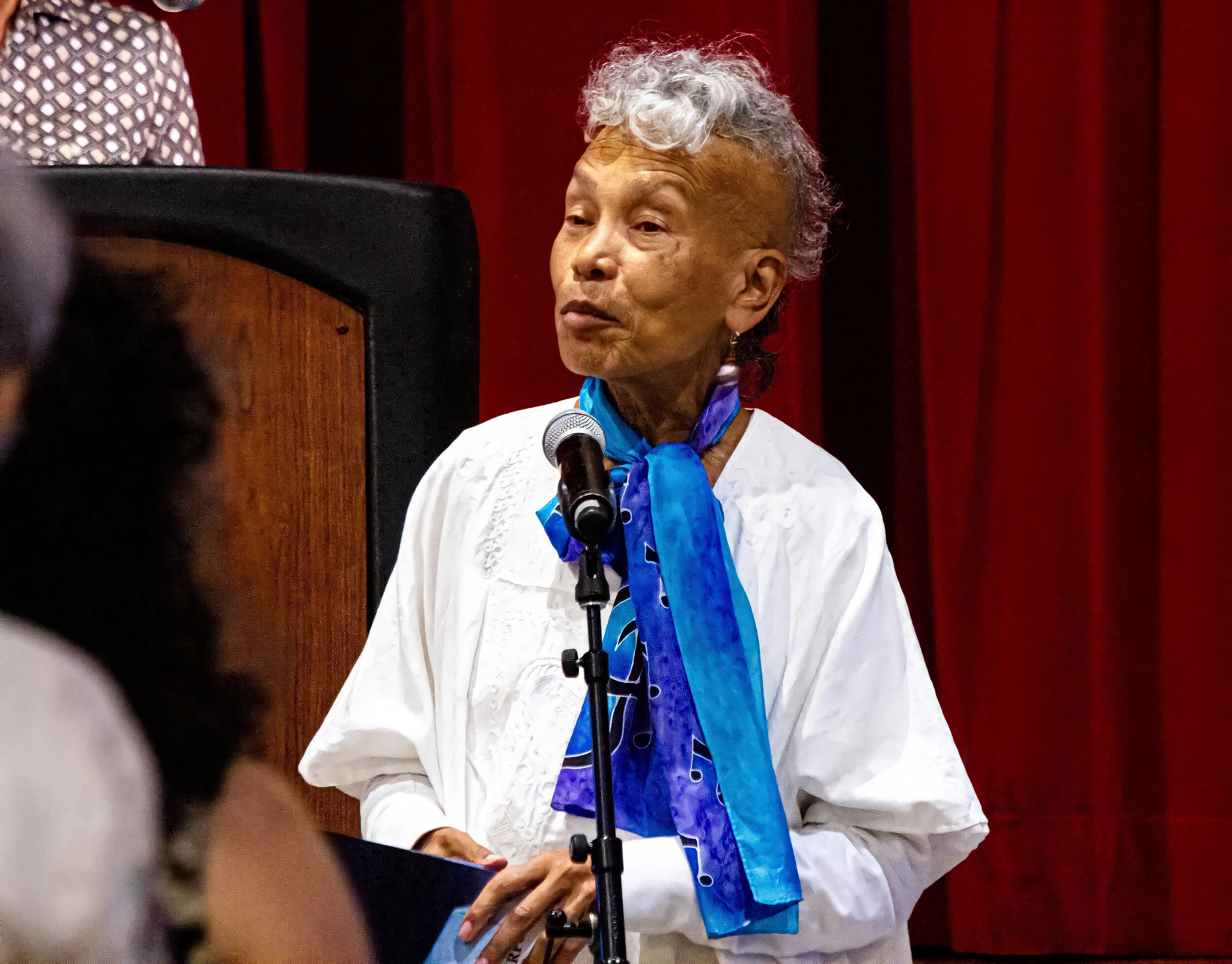 Older woman with gray curly hair and light brown skin wearing a white top and a blue scarf, sitting at a table with a microphone in front of her, against a red curtain background.